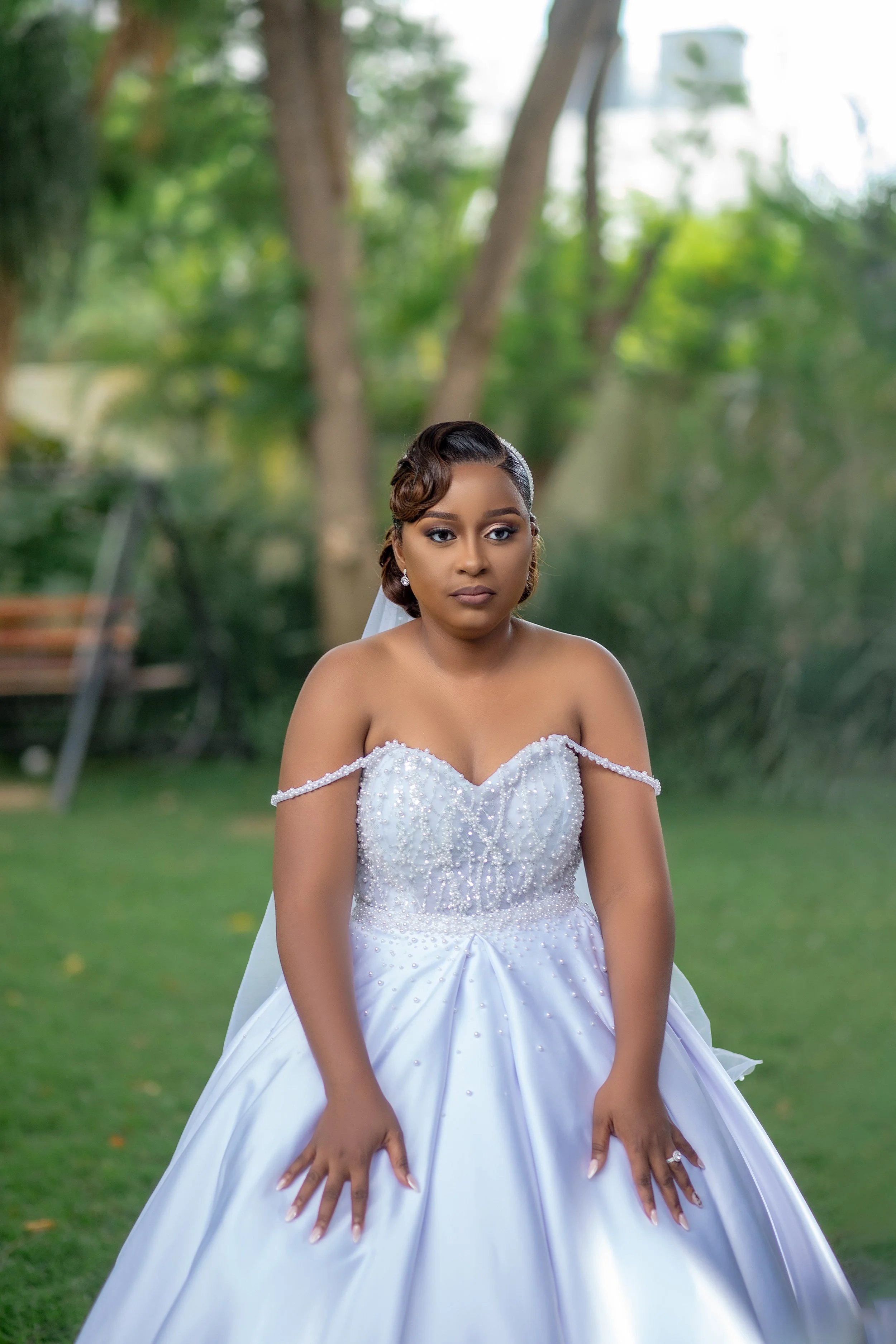 A bride wearing a white wedding gown with bead and pearl embellishments, standing outdoors with green trees and grass in the background.