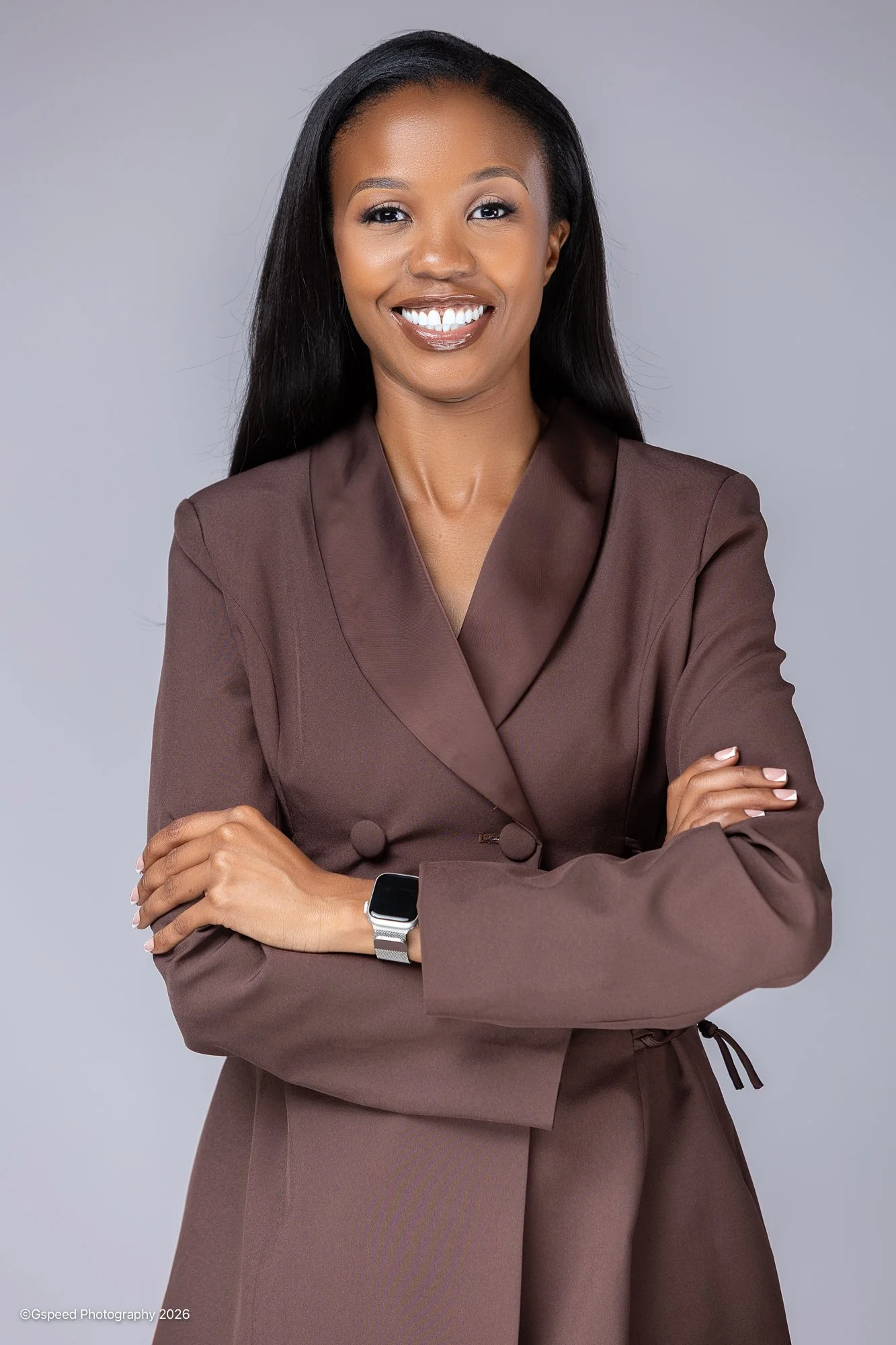 Portrait of a woman with long black hair in a brown blazer, smiling with crossed arms, wearing a smartwatch, against a plain gray background.