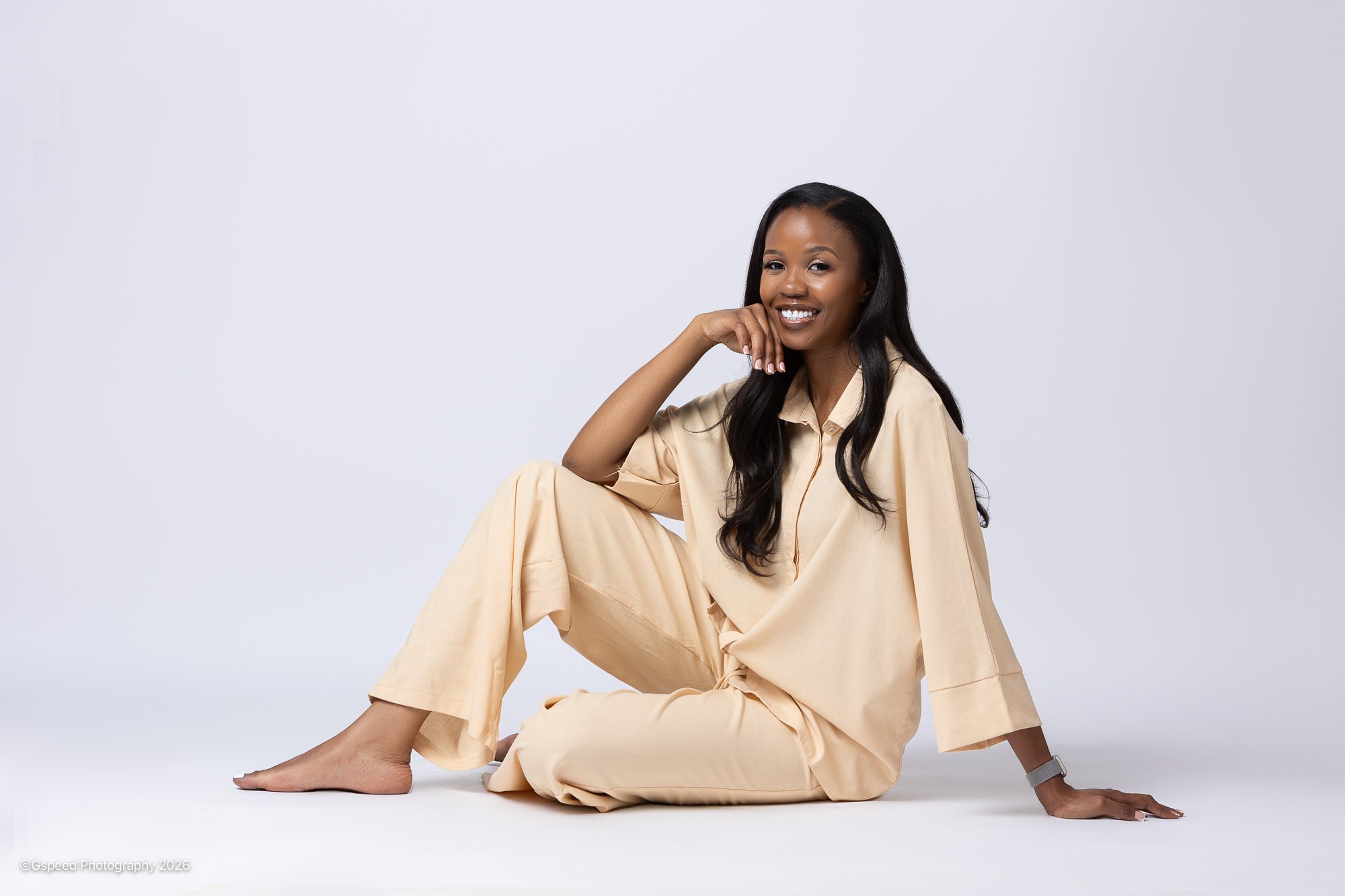 A woman with dark hair smiling, sitting on the floor with one leg bent and the other extended, wearing beige loose pajamas in front of a plain white background.