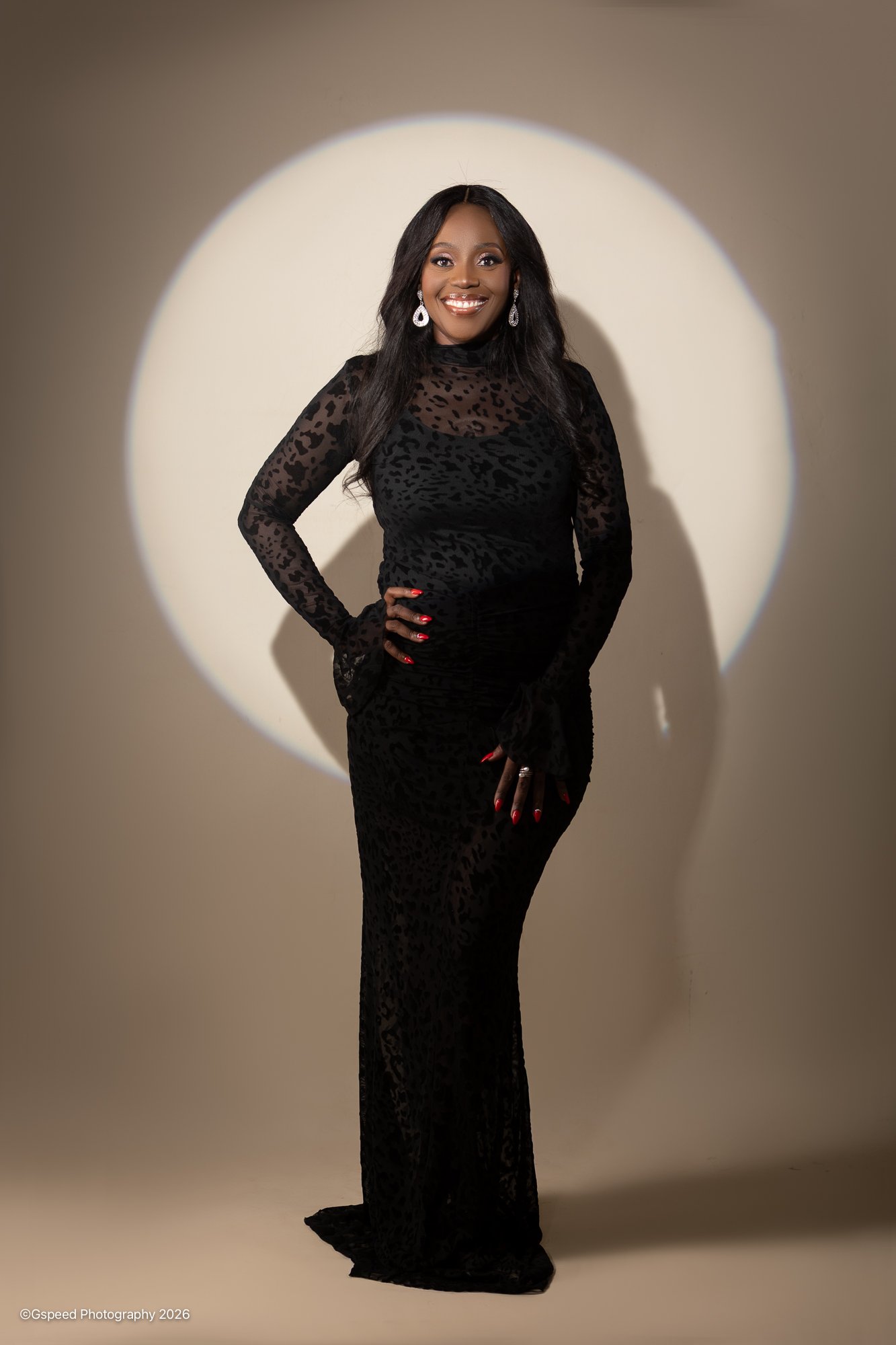 Portrait of a woman in a black leopard print gown standing against a neutral background with a spotlight creating a circular light effect behind her.