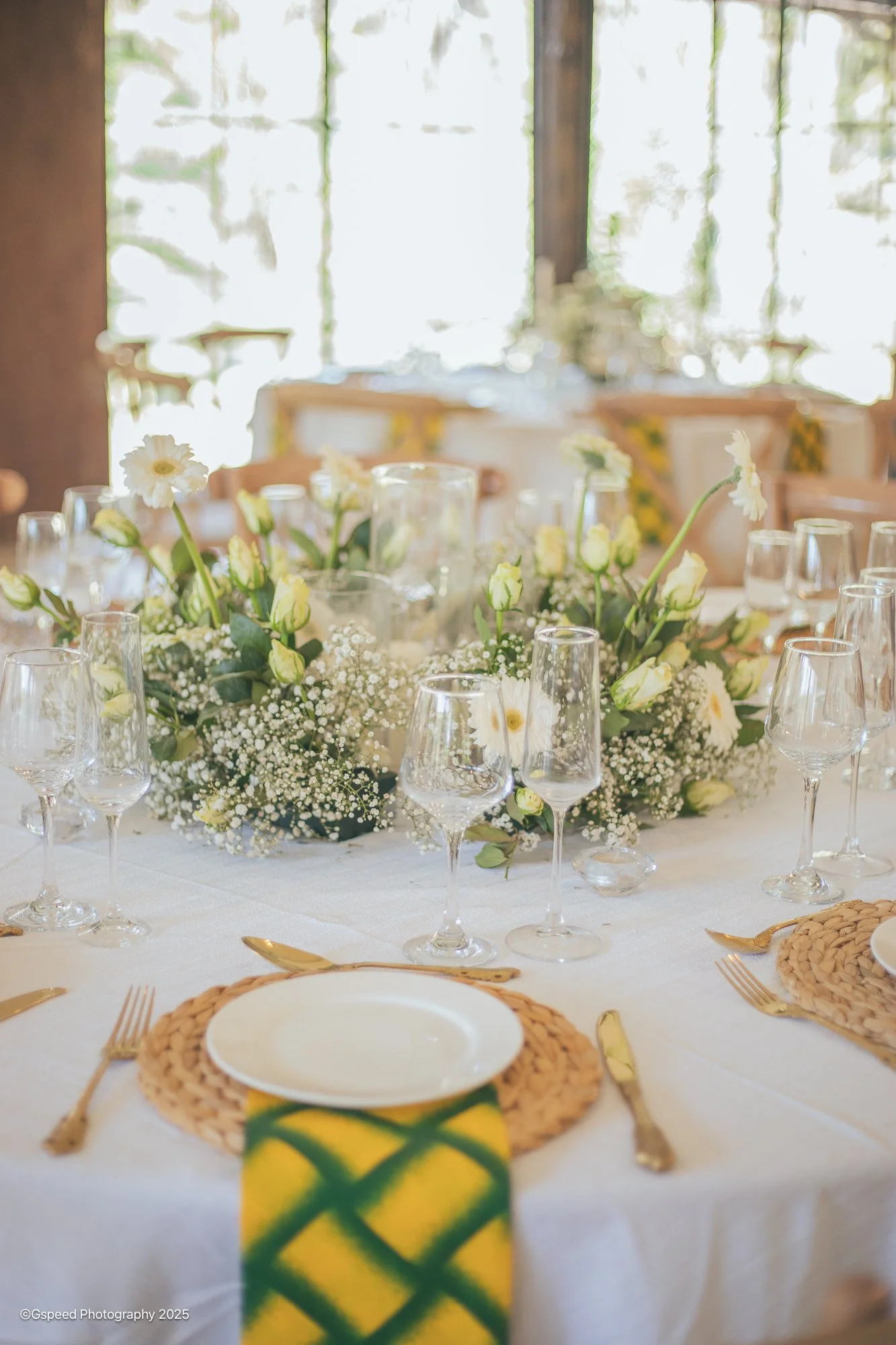 A beautifully decorated dining table with a floral centerpiece consisting of white flowers and green leaves, surrounded by glasses, gold cutlery, and woven placemats, set in a bright room with large windows allowing natural light.