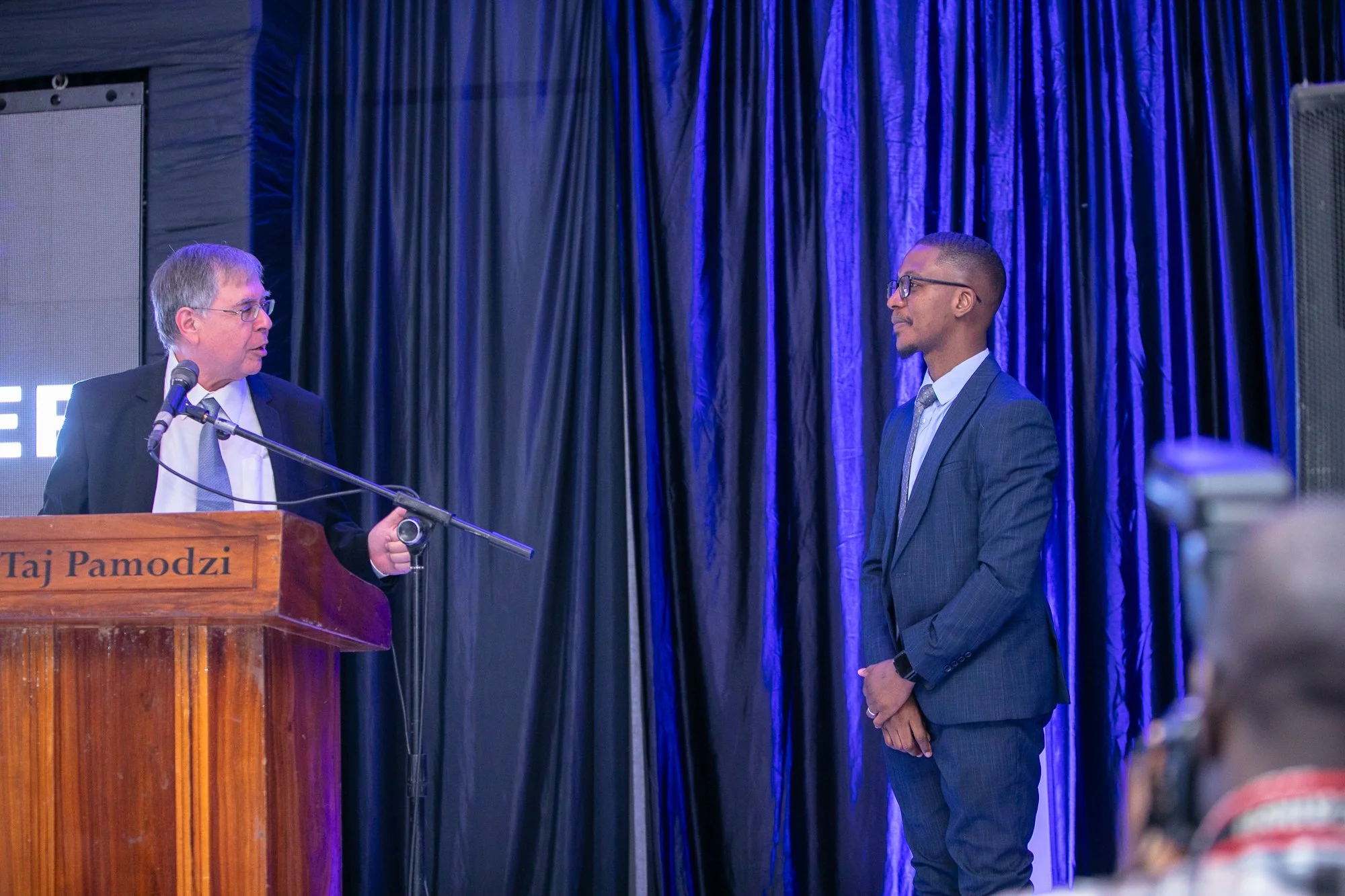 Two men are engaged in conversation on a stage with dark curtains. One man, wearing a dark suit and glasses, is speaking at a wooden podium labeled 'Taj Pamodzi.' The other man, dressed in a suit and glasses, is listening attentively. A microphone is