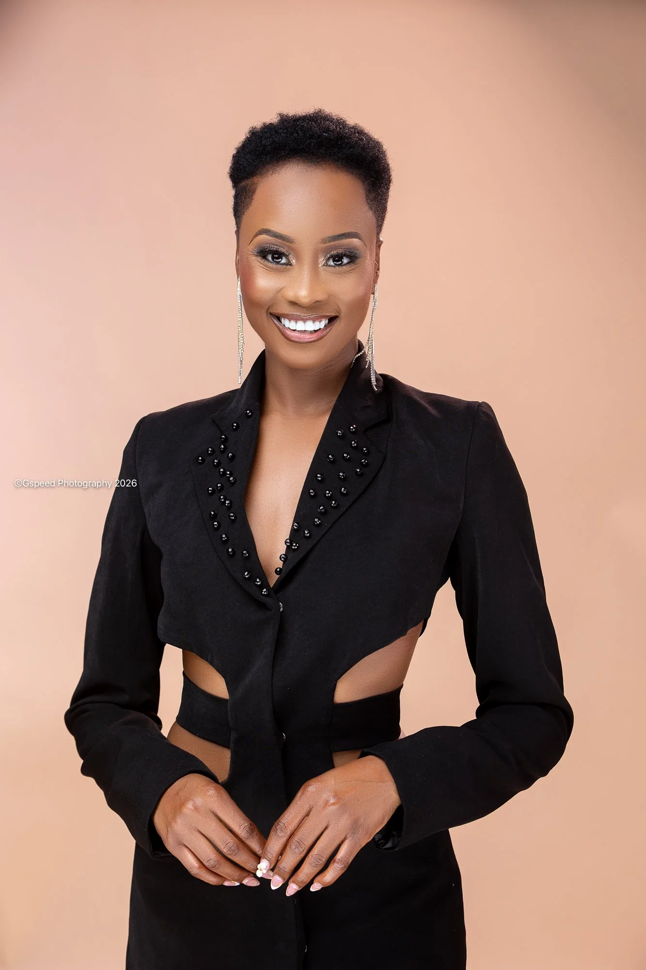 A woman with short black hair, wearing a black blazer with embellishments, smiling against a beige background.