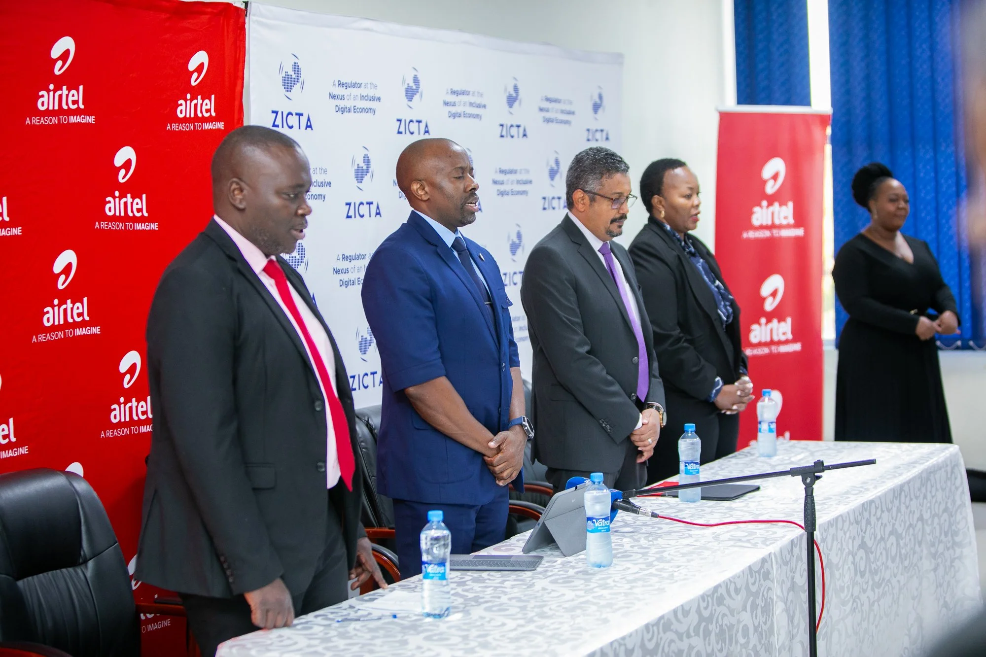 Five people in formal attire standing behind a table during a conference or panel discussion. There are banners with the logos and names of Airtel and ZICTA behind them. Water bottles are on the table, and a microphone is visible in the foreground.