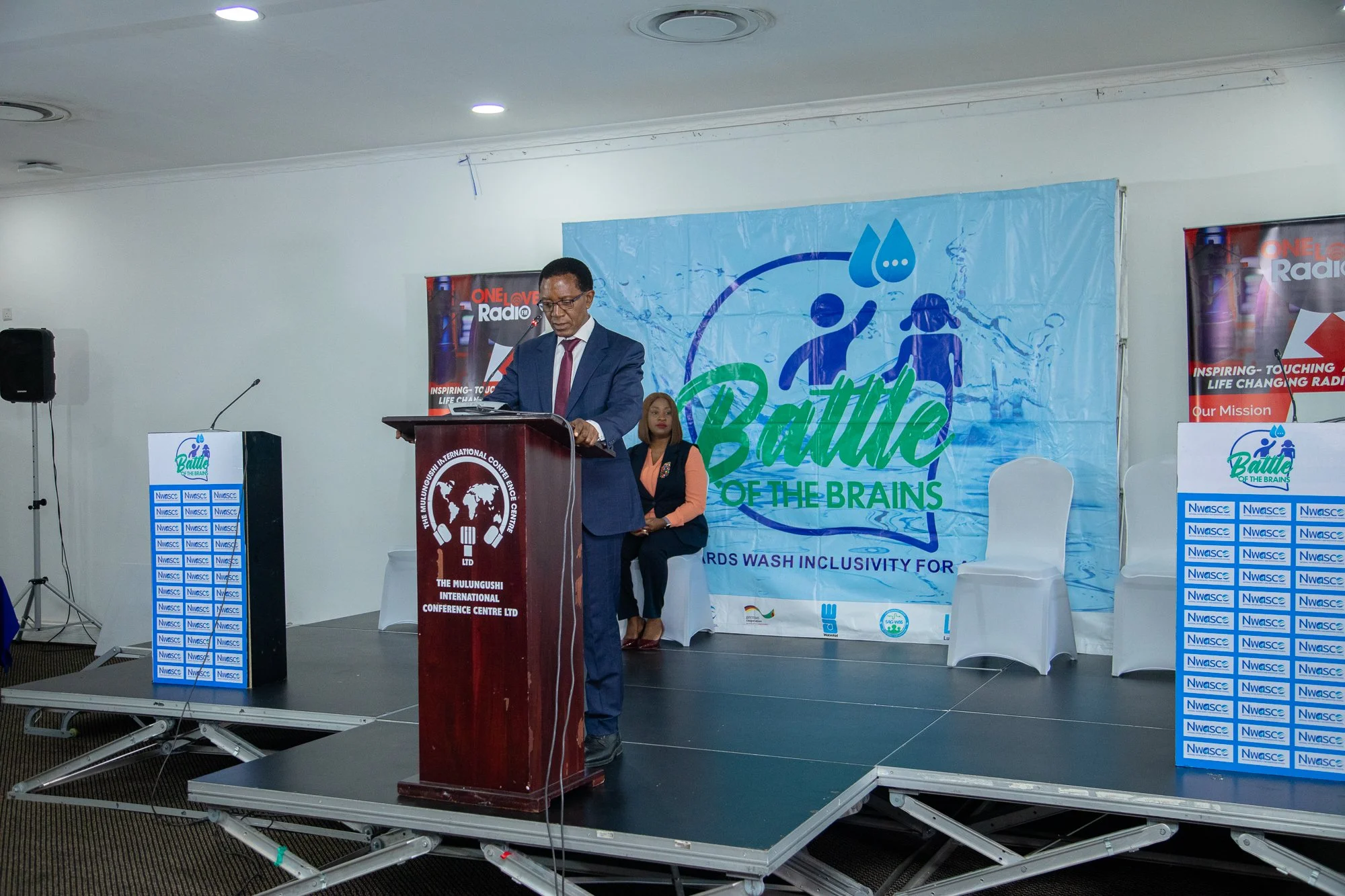 A man in a suit standing behind a podium at a conference, with a woman sitting beside him. The background has banners with the words 'Battle of the Brains' and 'Ngwache'. The room is set up for a presentation or speech.