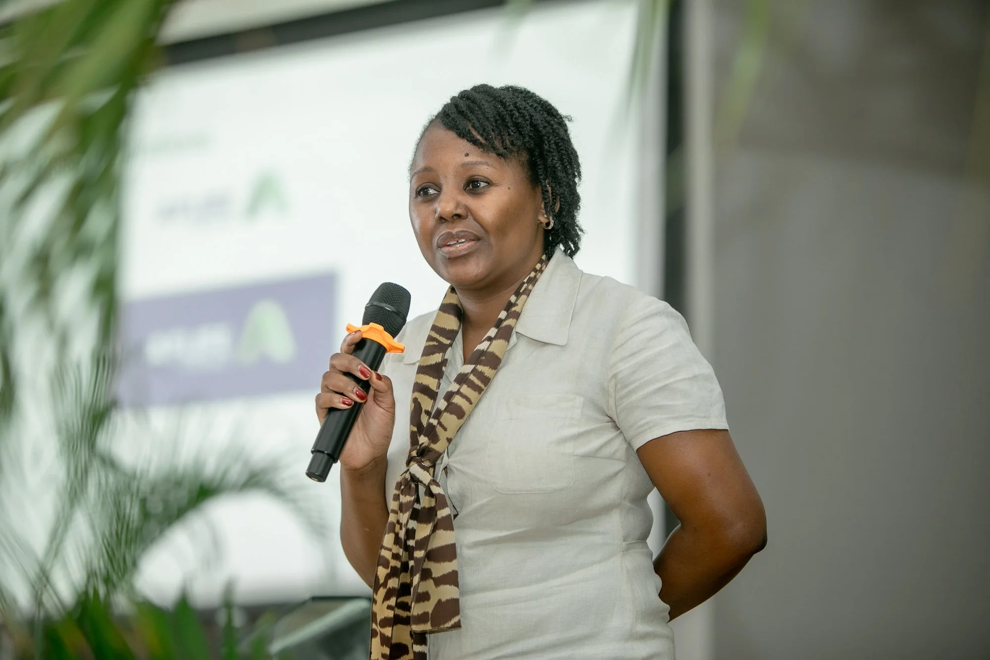 A woman holding a microphone speaking at an event, standing indoors with a blurred background.