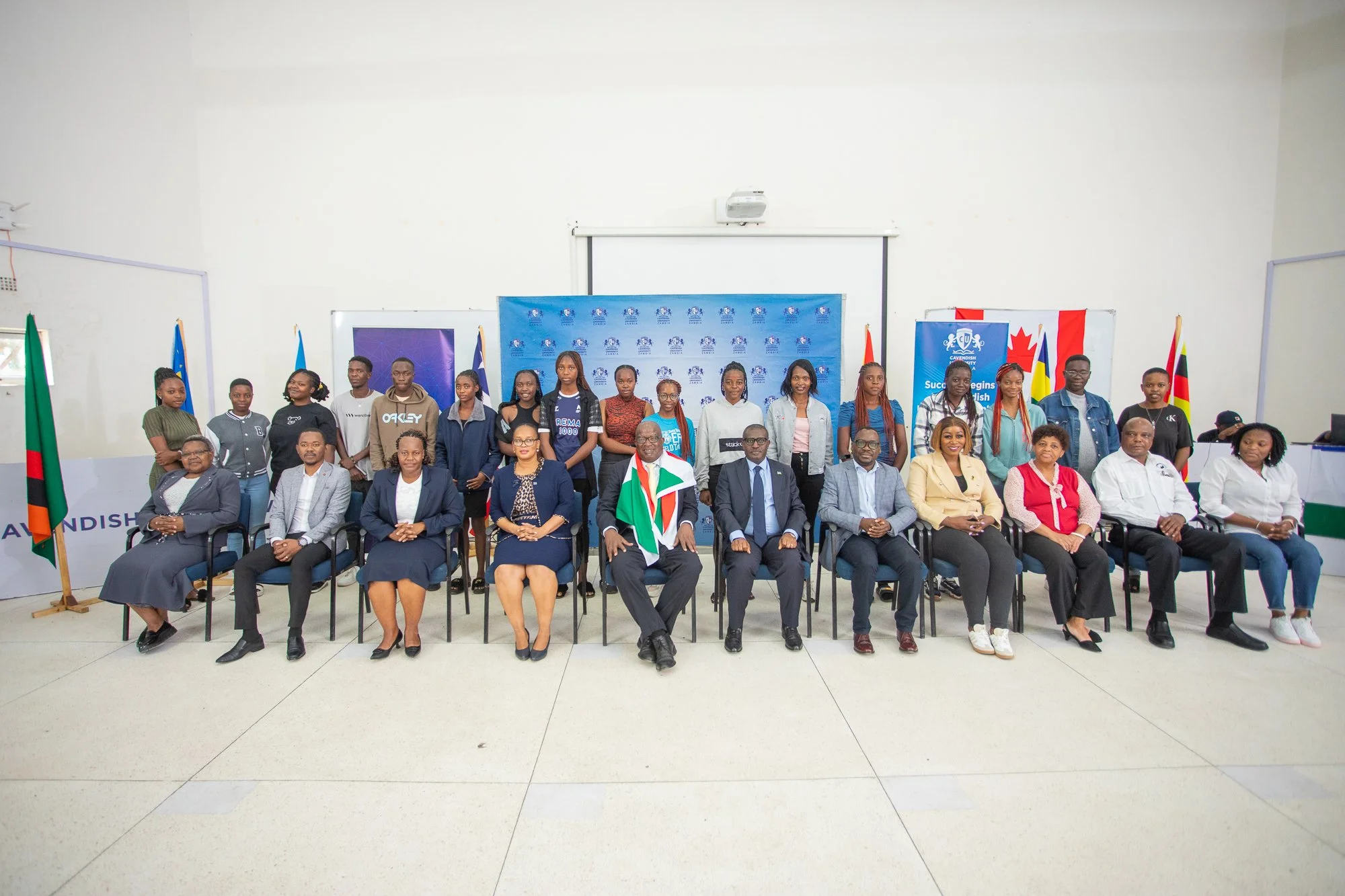 A group of people, including officials and young adults, posing for a photo in a room decorated with flags and banners, some seated in front and others standing behind.