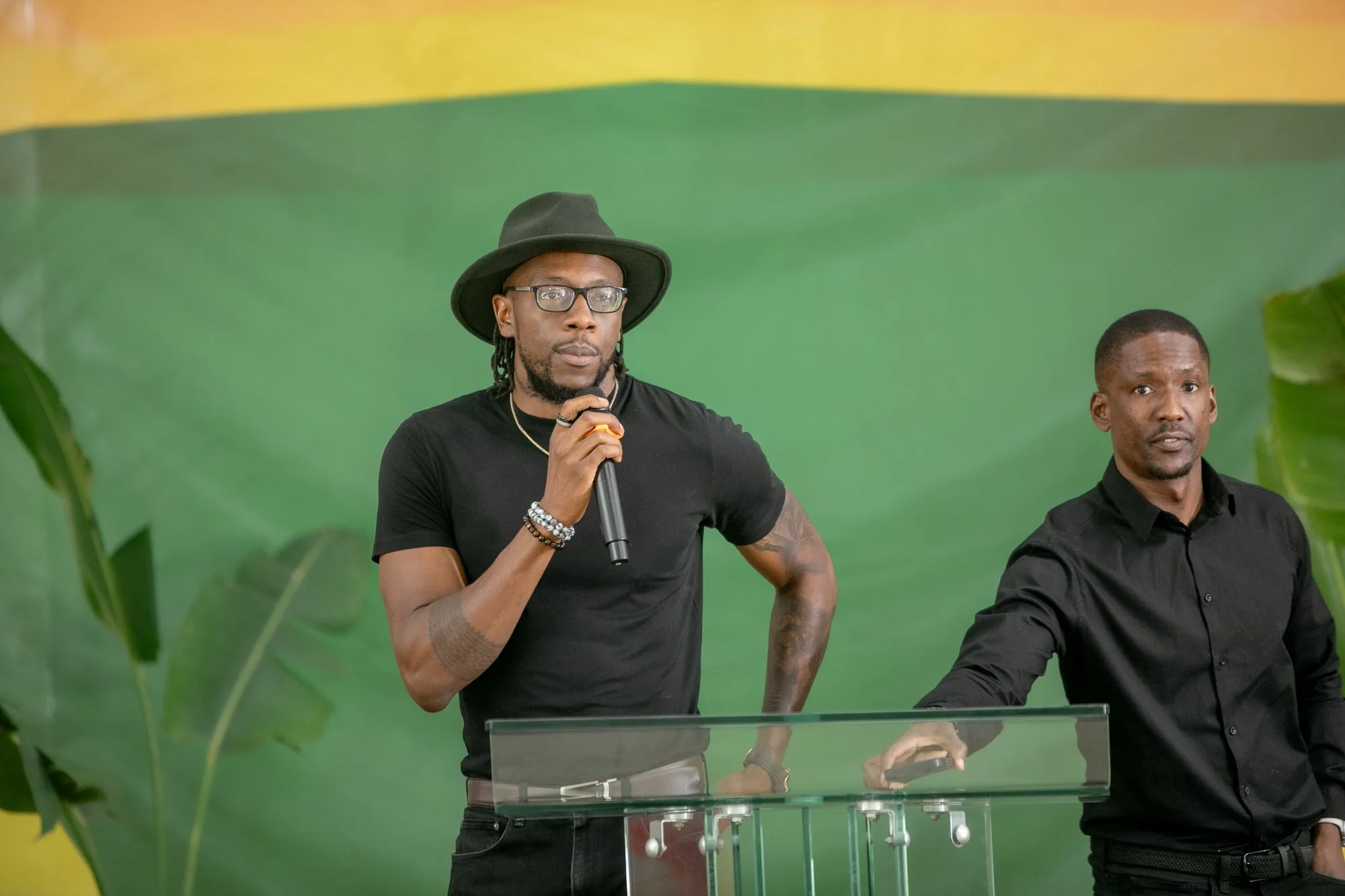 Two men stand behind a transparent podium, one speaking into a microphone, dressed in a black shirt and wearing glasses and a black hat, with a green background.