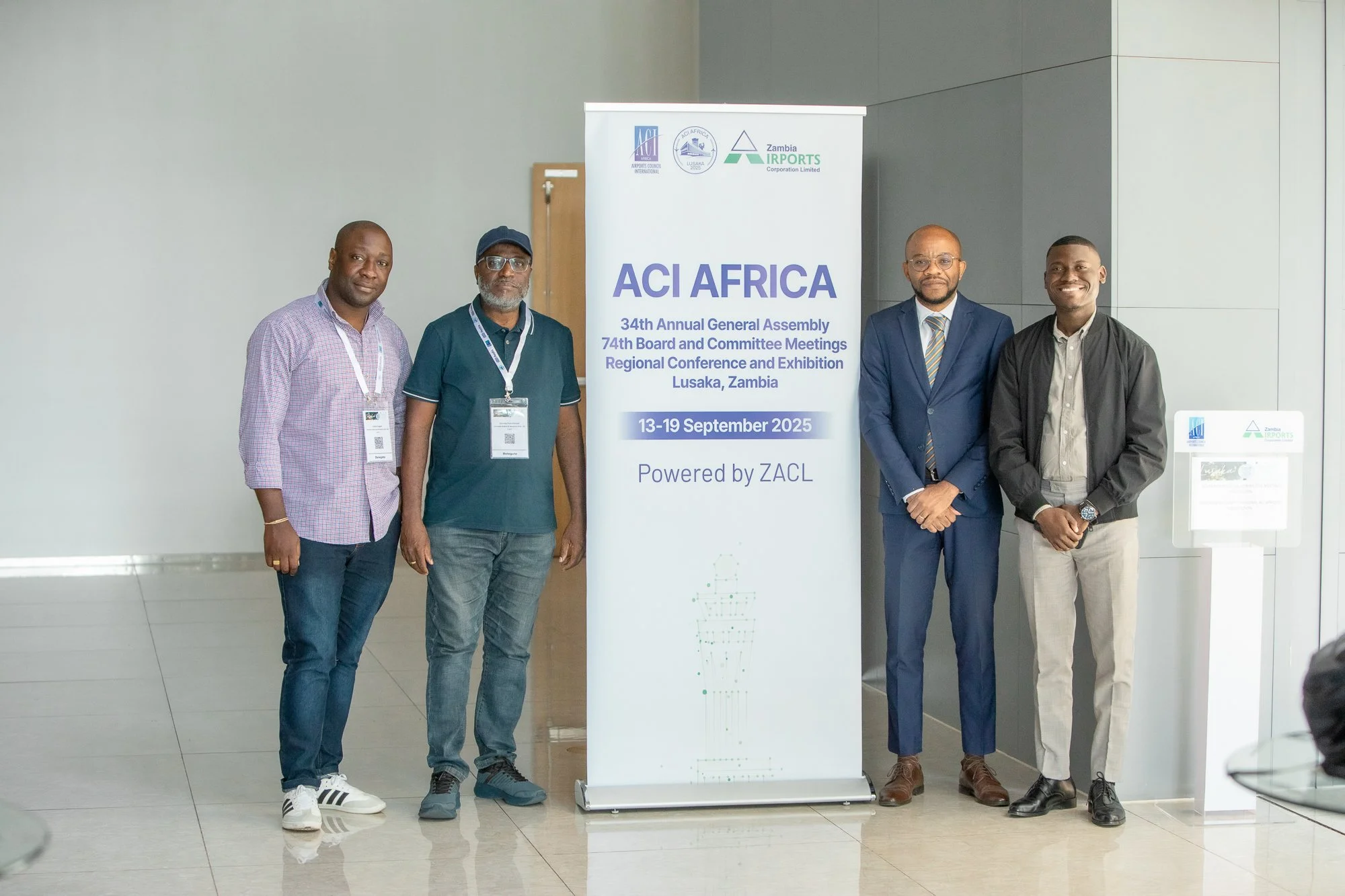 Four men standing beside a white banner with logos and text about ACI Africa annual general assembly, in Lusaka, Zambia, September 2025.