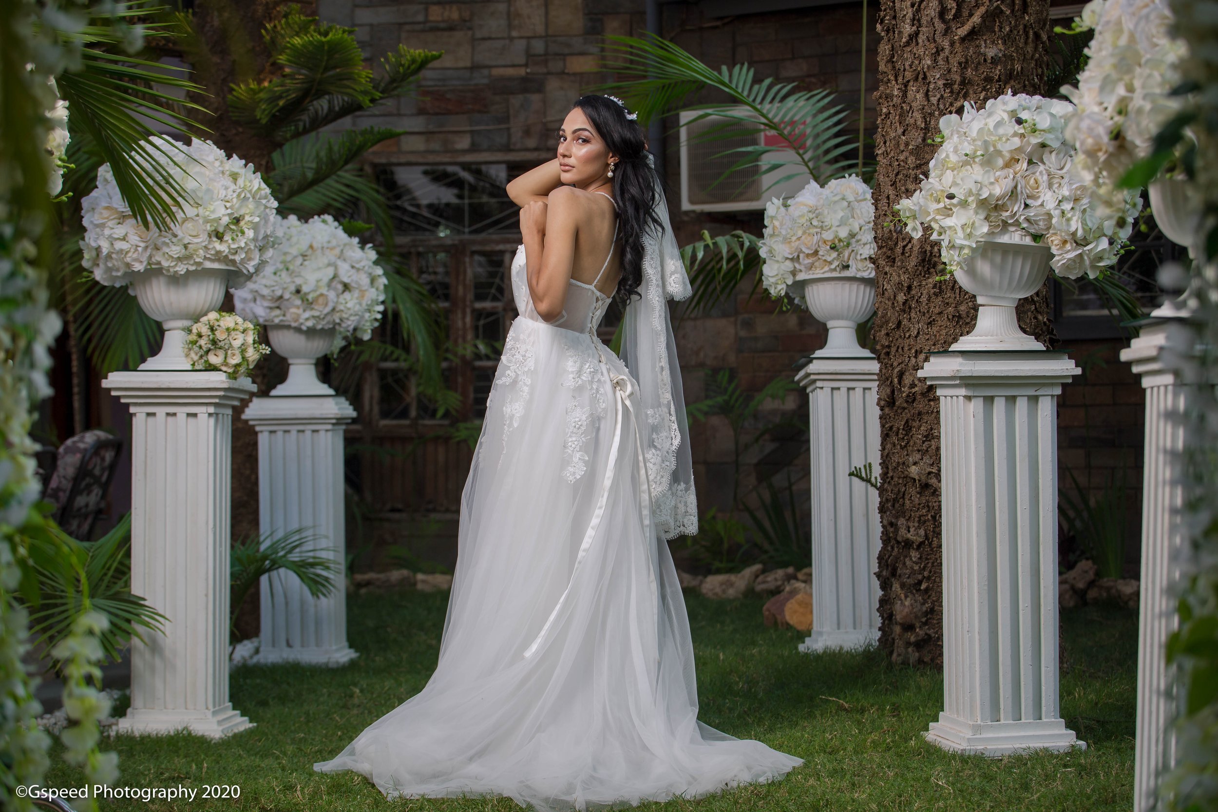 A bride in a white wedding dress with lace details, standing outdoors among white floral arrangements on pillars and green foliage, looking over her shoulder.