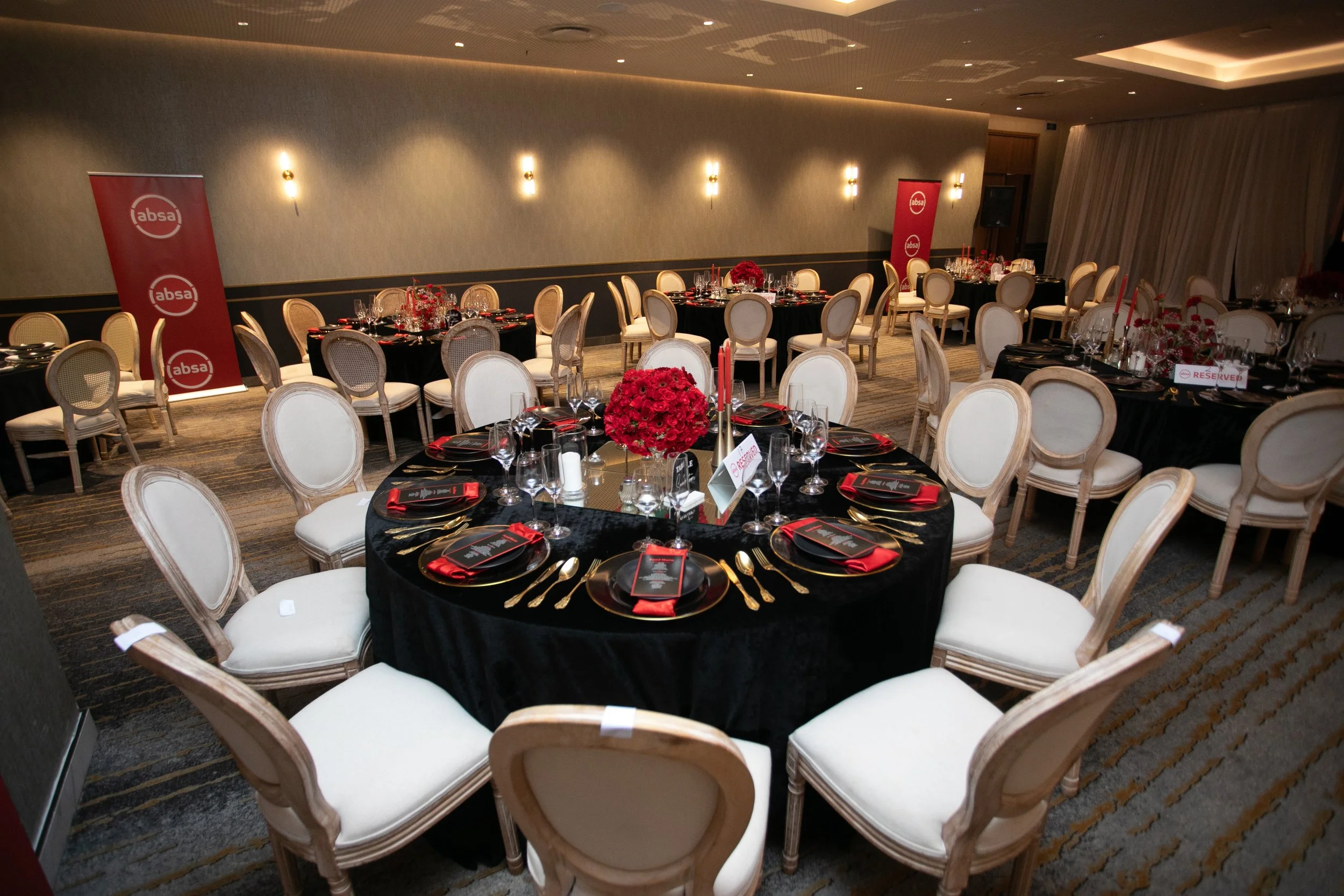 Formal dining area decorated with red and black accents, featuring round tables with black tablecloths, white chairs, and floral centerpieces, set for an event, with Absa banners and reserved signs.