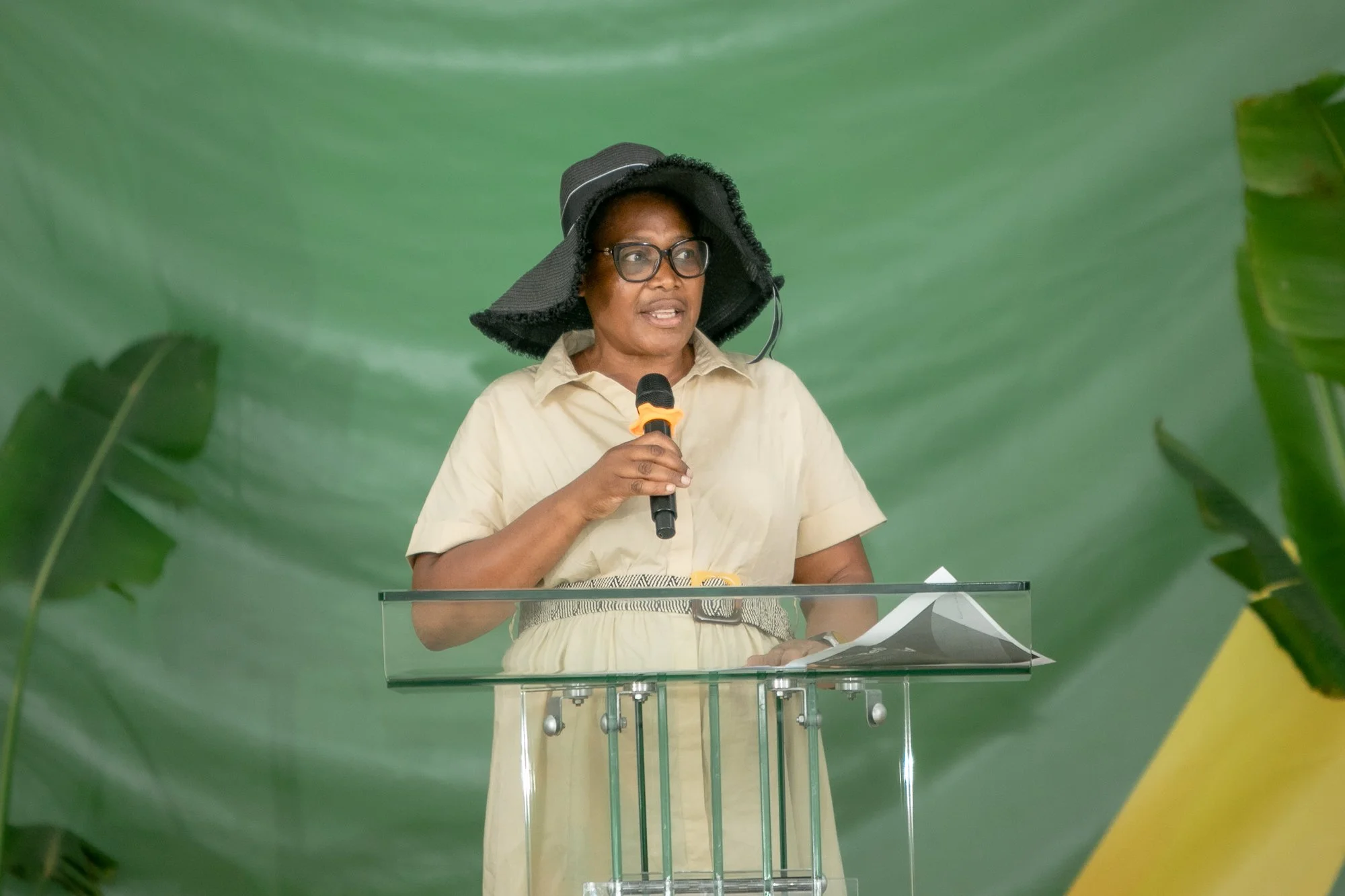 A woman wearing glasses and a wide-brimmed hat is speaking into a microphone at a clear podium in front of a green leafy background.