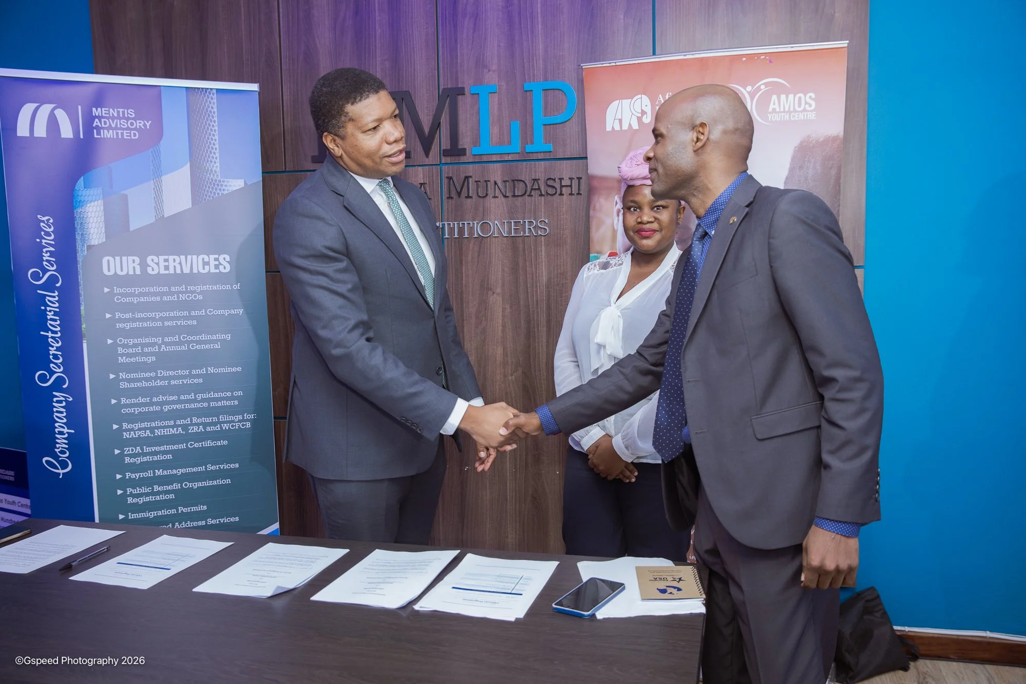 Two men in suits shaking hands at an event, with a woman in traditional attire standing behind them. Behind them are banners with text related to corporate services and organizations, and documents are laid out on a table in front of them.