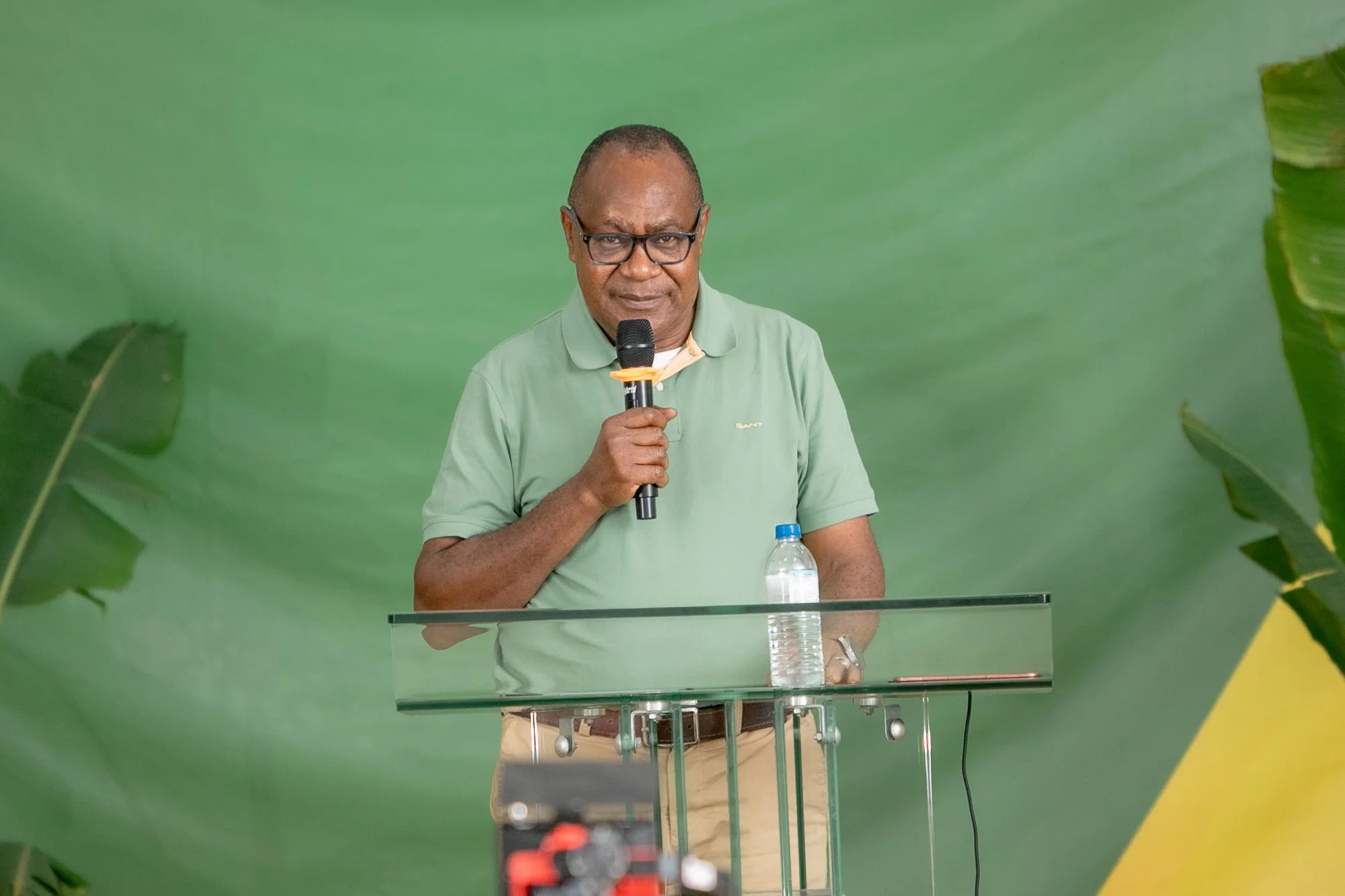 A man in glasses and a green polo shirt is giving a speech at a podium, holding a microphone in his right hand with a water bottle on the podium, against a green background with large leaves visible on either side.