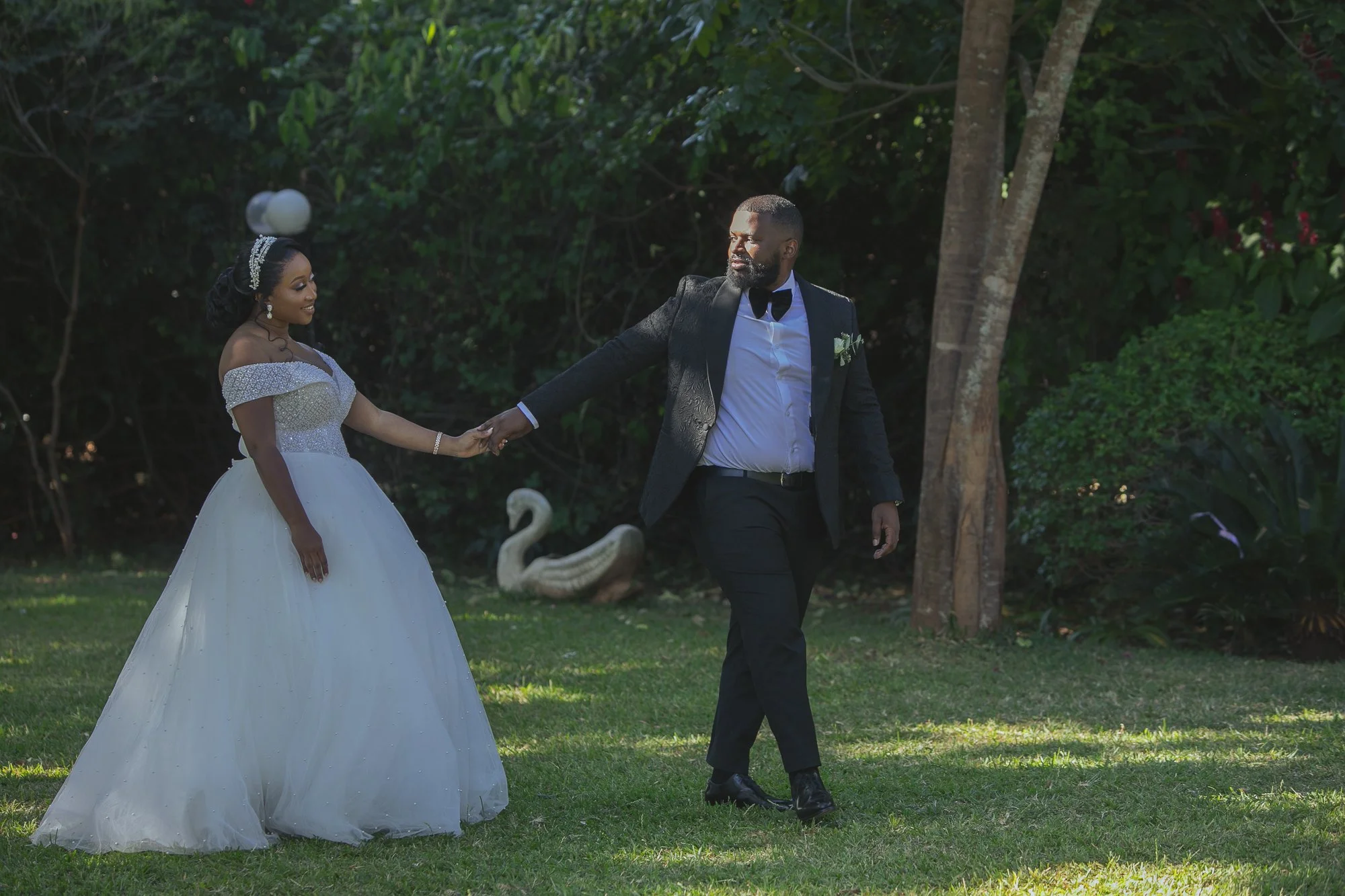 A bride and groom holding hands outdoors in a garden with lush greenery, trees, and decorative swan statues, celebrating their wedding day.