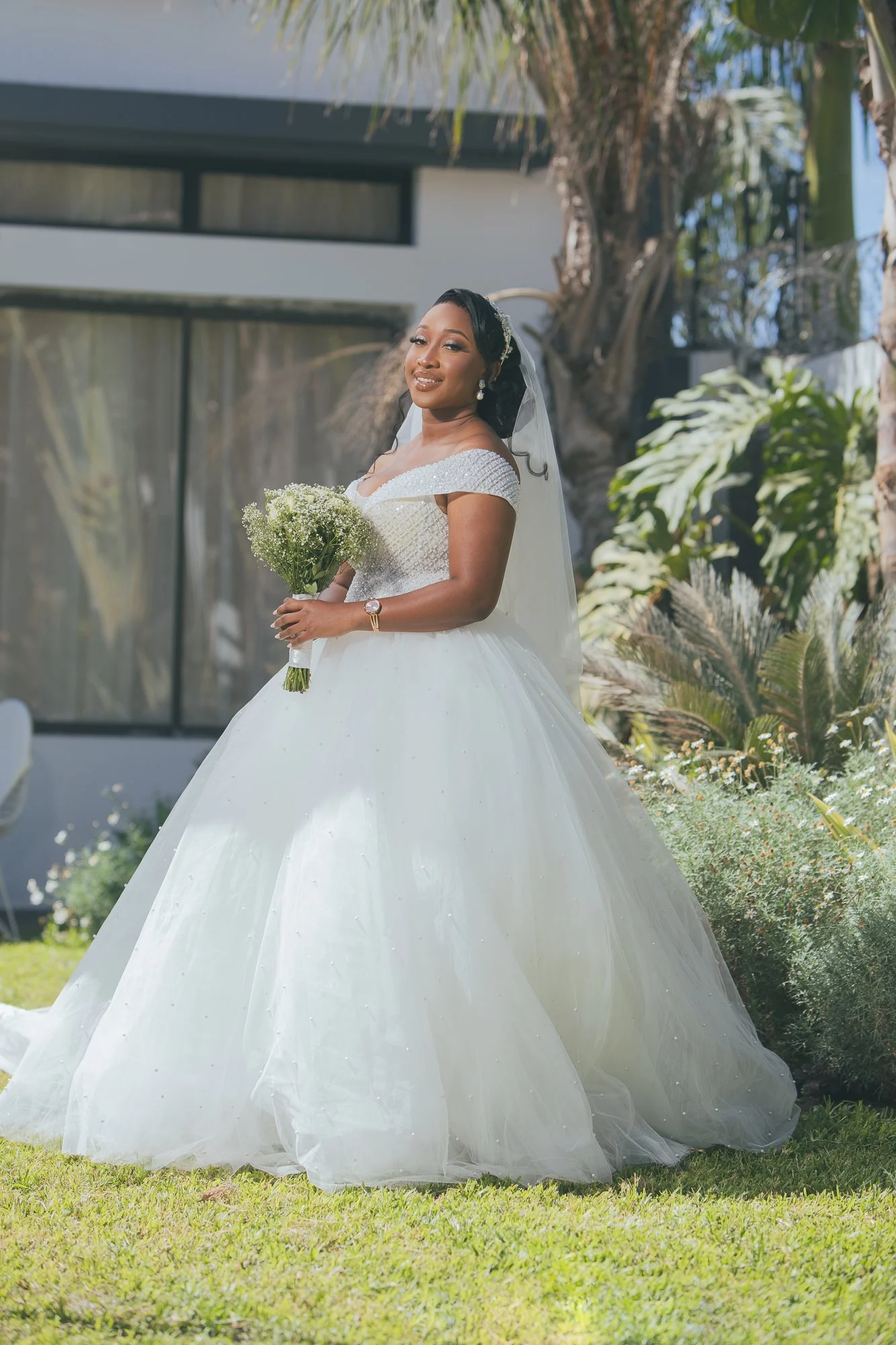 A bride in a white wedding gown holding a bouquet of white flowers, standing outdoors with greenery and trees in the background.