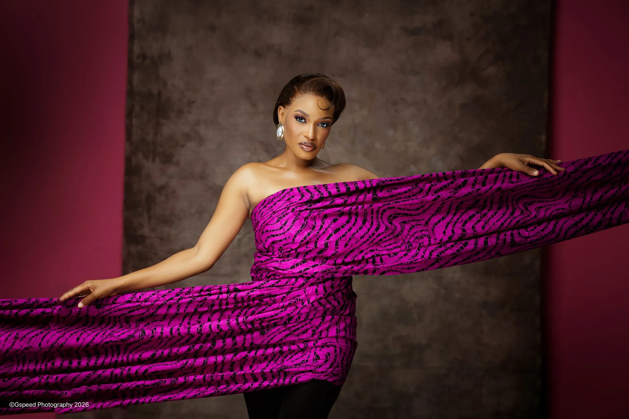 A woman with styled hair, makeup, and earrings holds a flowing piece of pink and black patterned fabric in front of a textured brown background with pink curtains on sides.