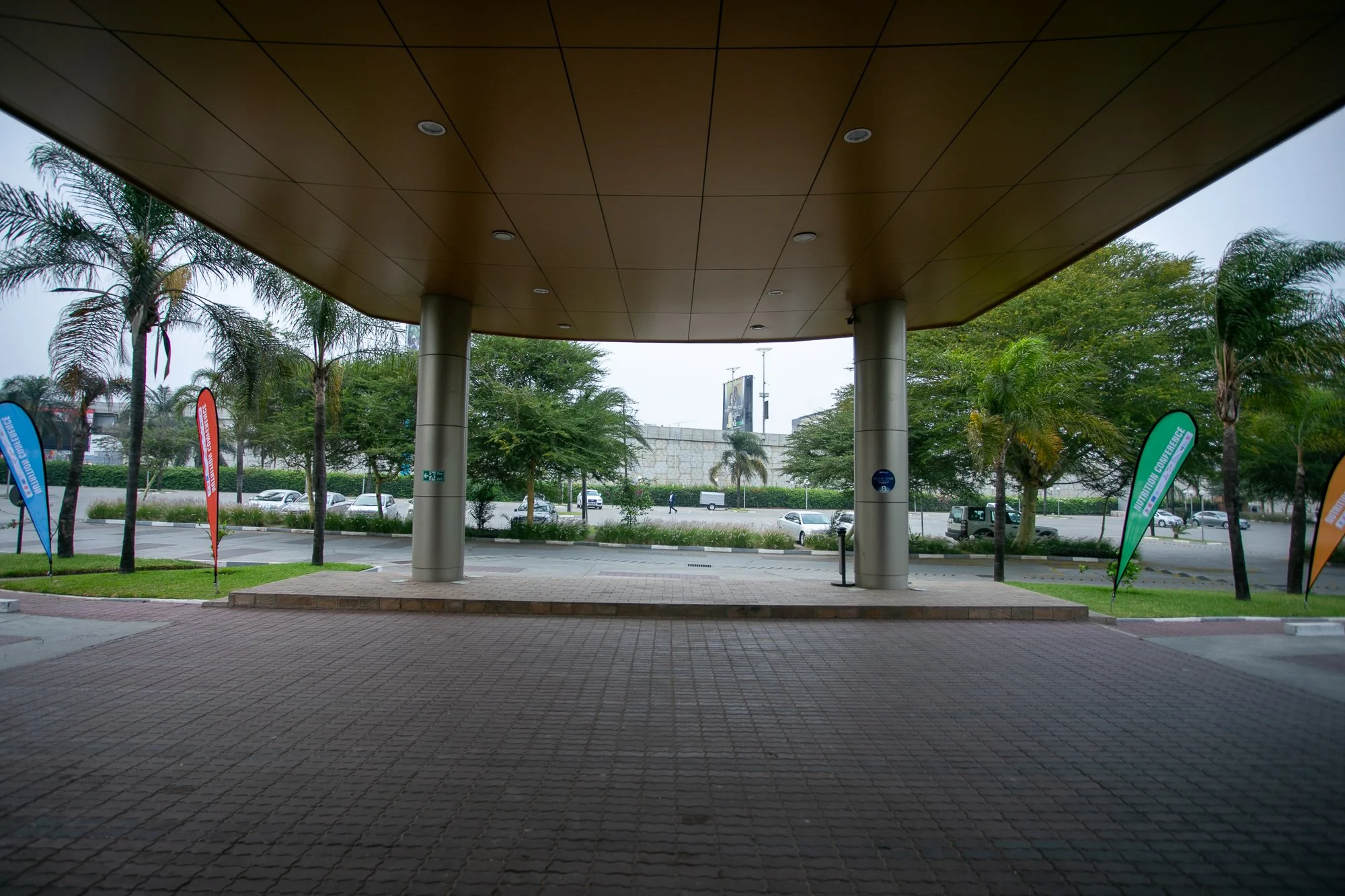 An outdoor scene with a large, overhanging brown roof supported by white columns. The area underneath the roof has a tiled floor and leads to a parking lot with several cars. In the background, there are palm trees, green bushes, and a street with mo