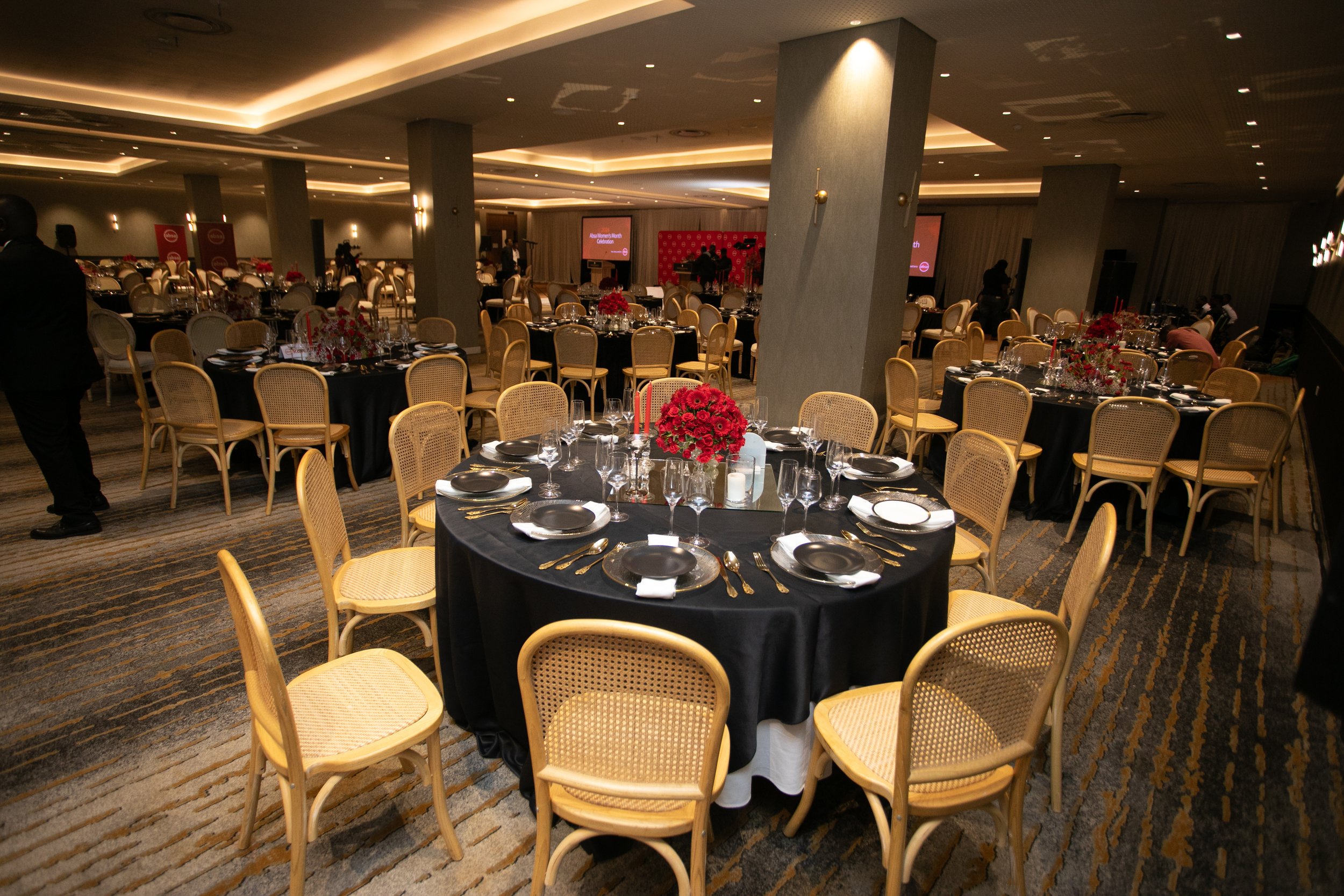 Banquet hall with round tables decorated with red floral centerpieces, black tablecloths, and place settings, ready for an event, with a stage and screens in the background.