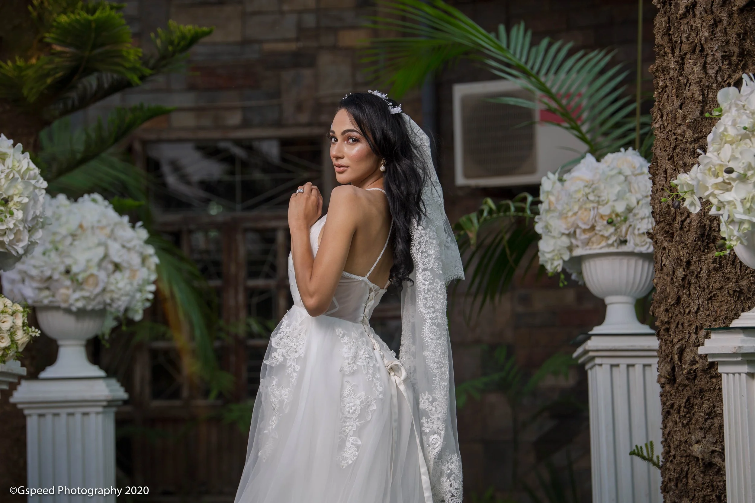 A bride in a white lace wedding dress with long dark hair, standing outdoors among white floral arrangements and green tropical plants, with a brick wall background.
