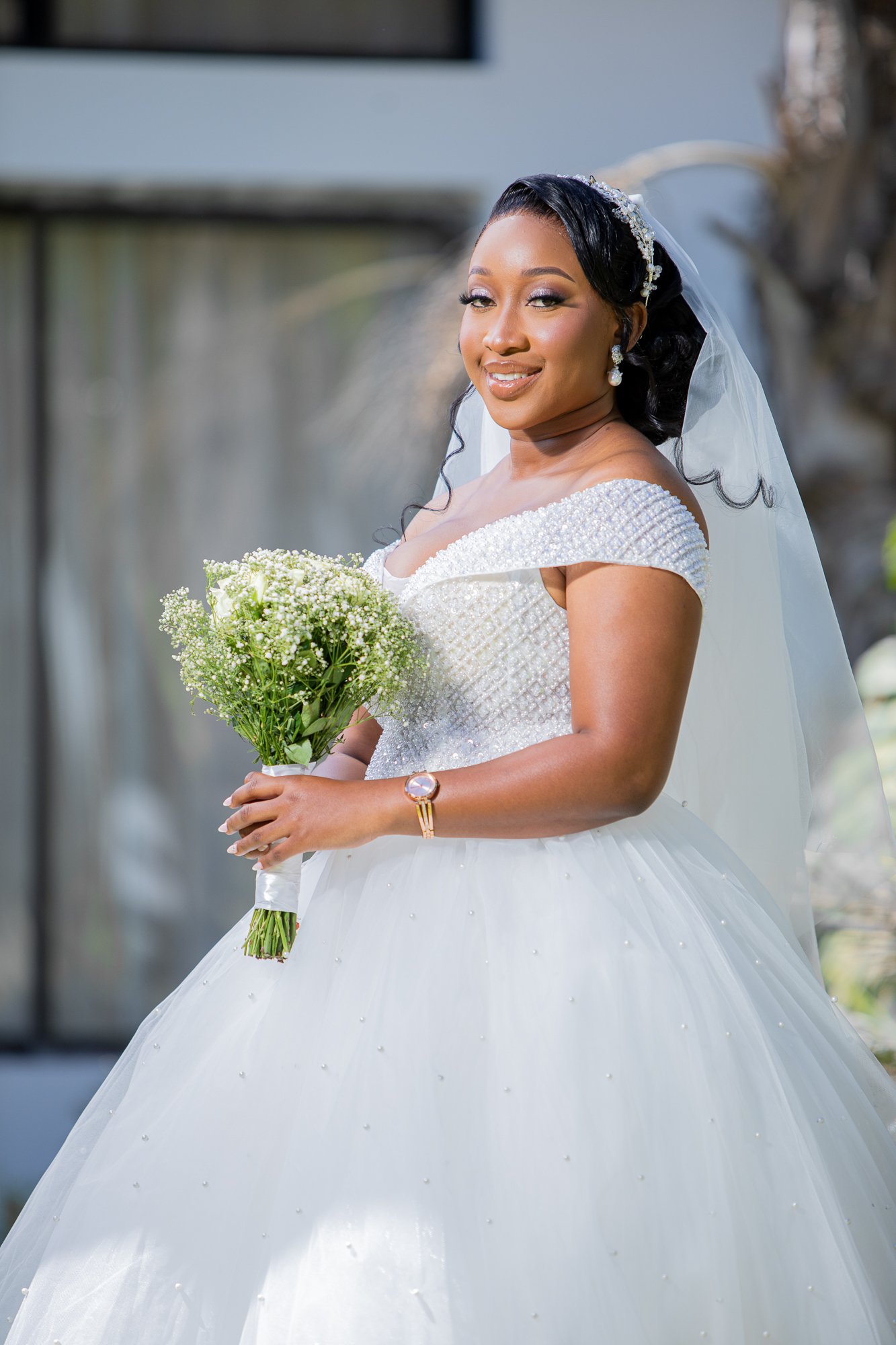 A smiling bride in a white wedding dress holding a bouquet of flowers, standing outdoors.