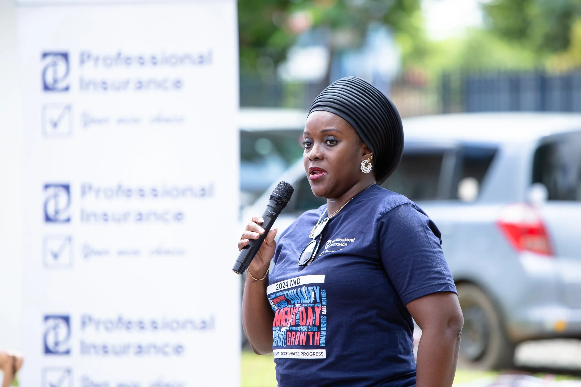 African American woman wearing a navy blue shirt with text, holding a microphone, speaking at an outdoor event with a blurred sign in the background.