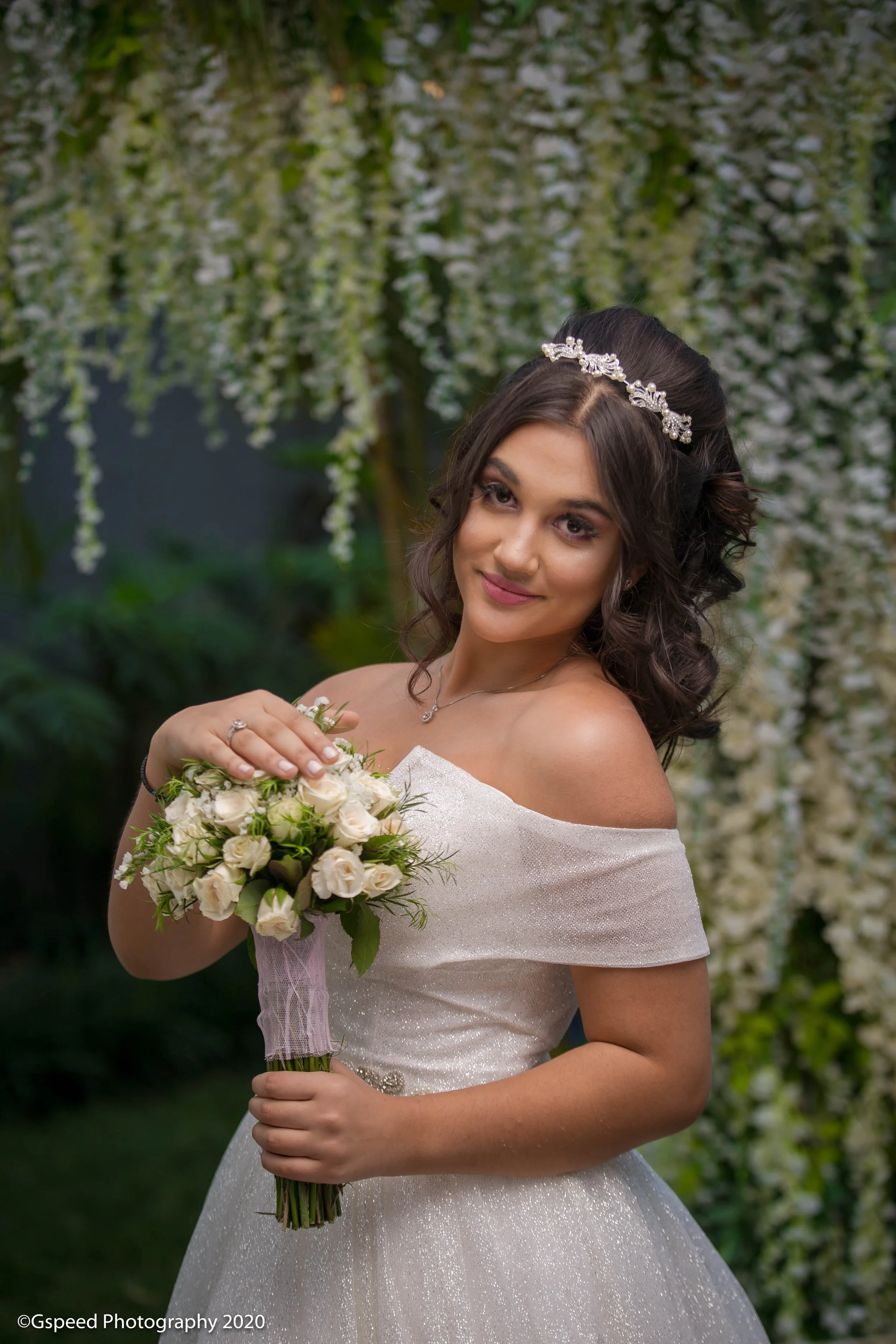 A bride holding a bouquet of white roses and greenery, wearing a white off-shoulder wedding gown with a sparkly texture, and a silver headband with floral design, standing in front of a backdrop of hanging white flowers.