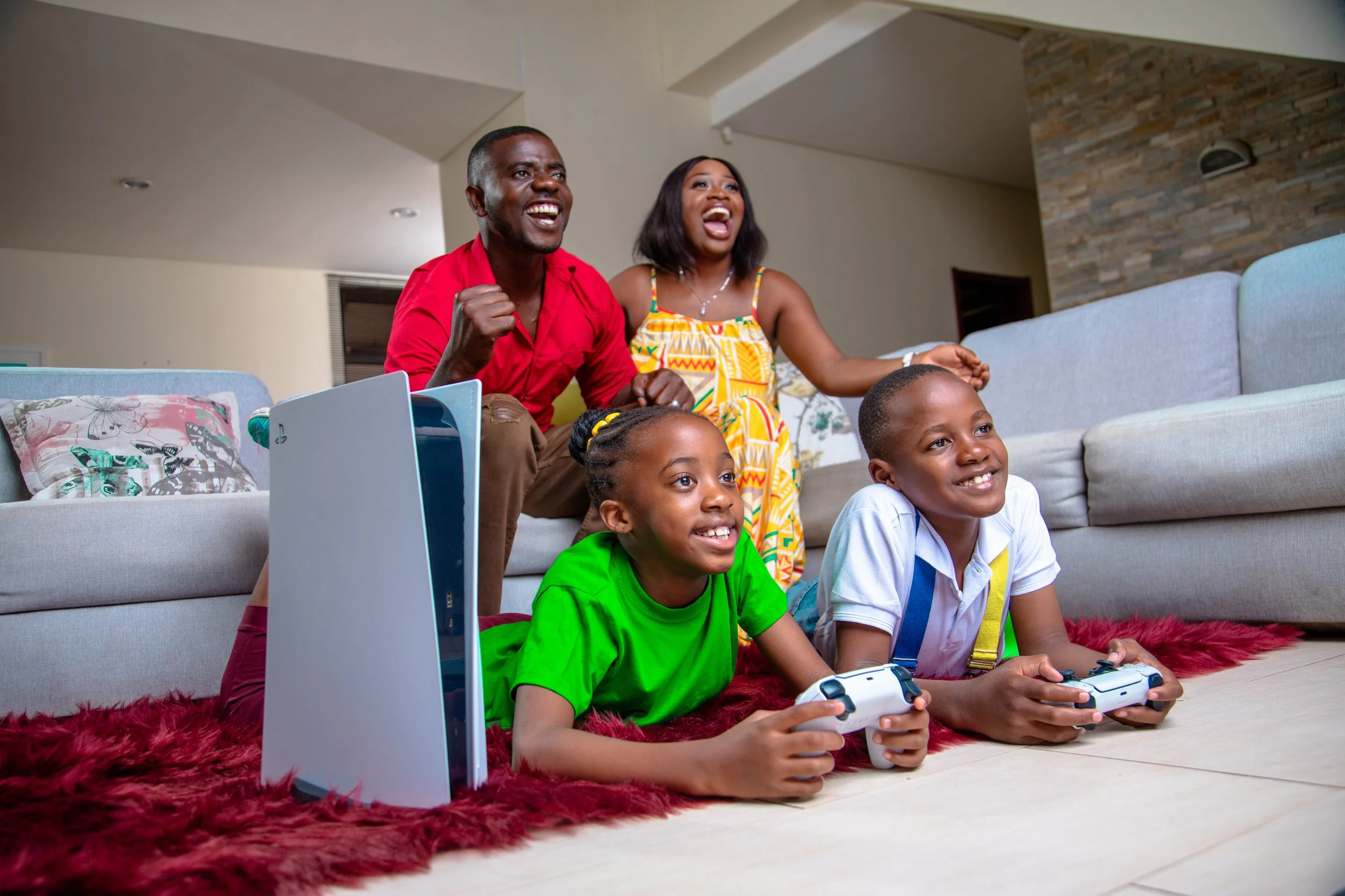 Family of four, two children and two adults, sitting on plush red rug on floor, playing video games, in living room, smiling and enjoying each other's company.