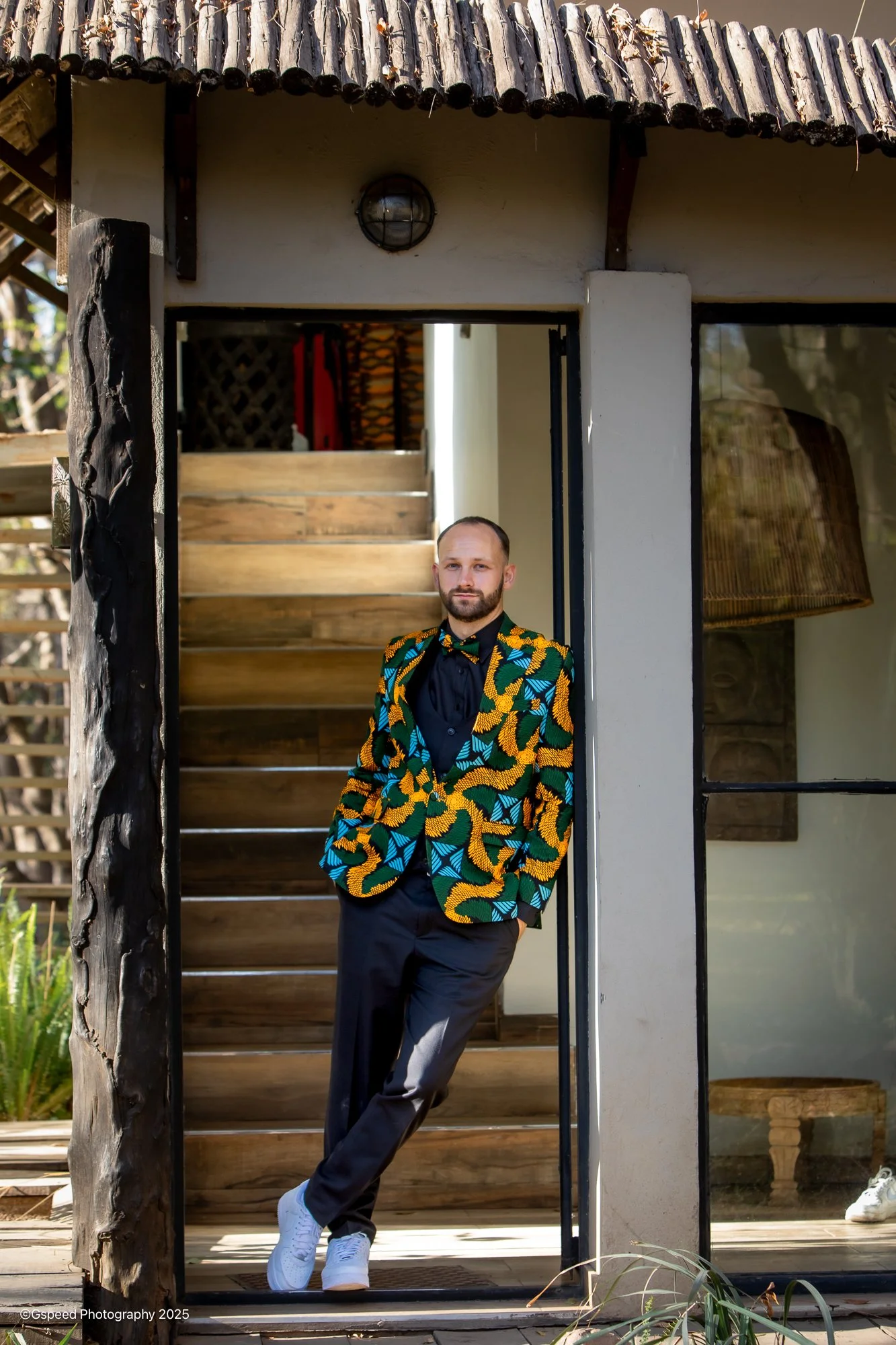 A man dressed in a vibrant, multicolored patterned blazer, black shirt, and black pants, leaning casually with one hand in his pocket in front of a modern building entrance with wooden stairs and a small outdoor porch.