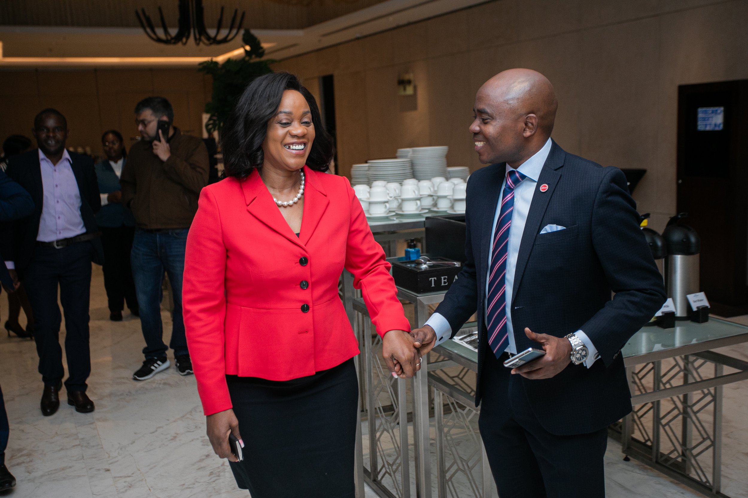 Two people shaking hands, a woman wearing a red blazer and a pearl necklace, and a man wearing a dark suit, striped tie, and a badge, smiling at each other in a professional setting with other people in the background.