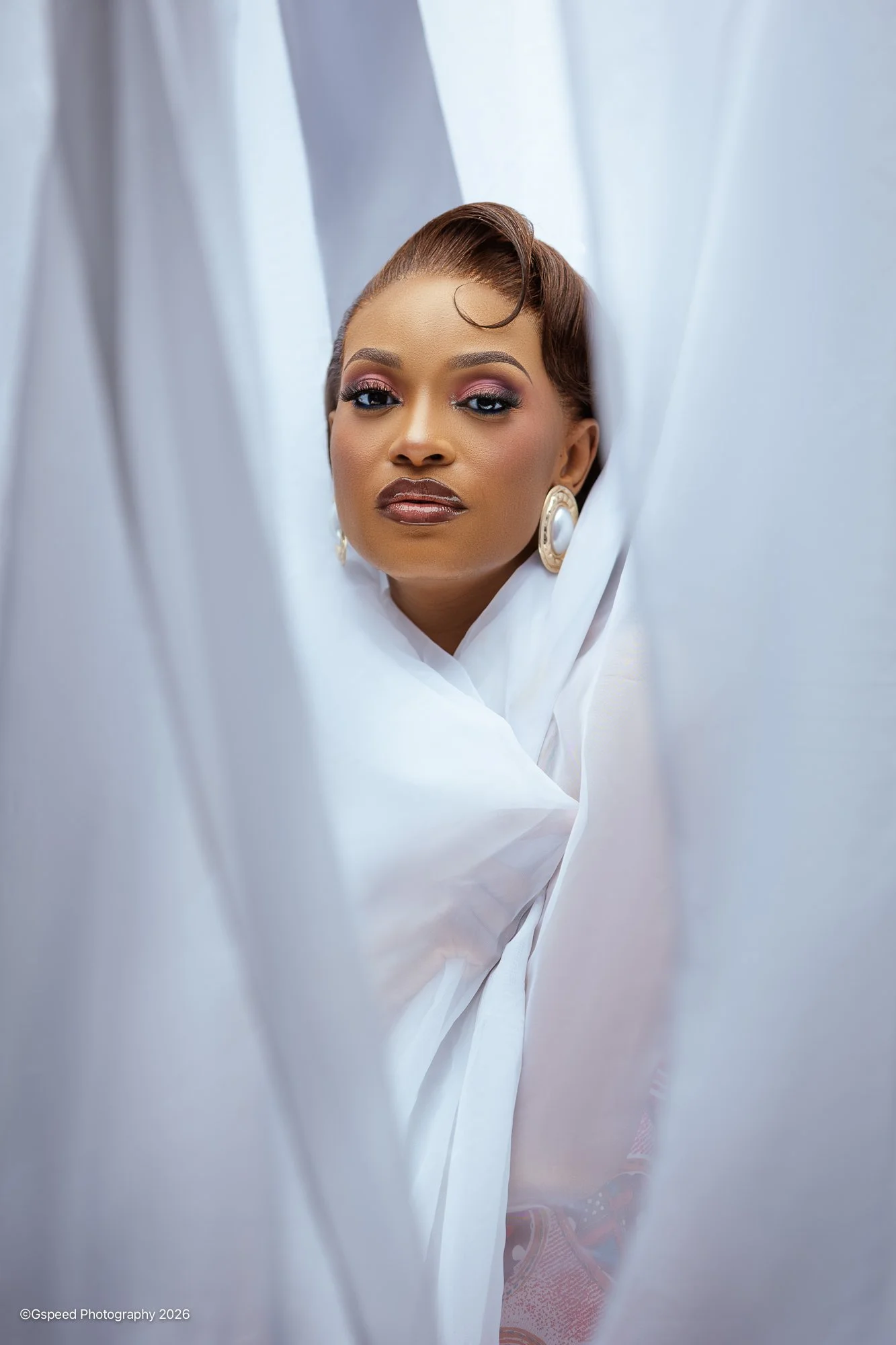 A woman with short brown hair and large earrings looking through white curtains.