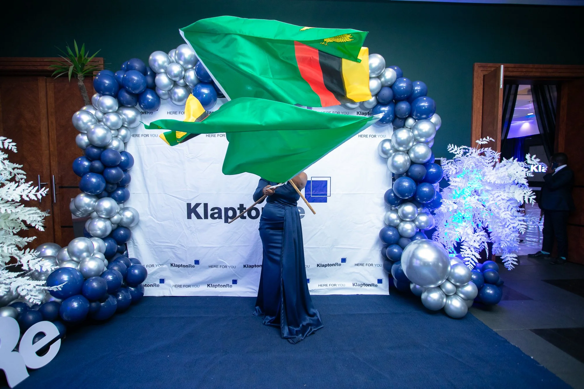 Person holding two Zimbabwean flags at a formal event with blue and silver balloon decorations and a white backdrop with the KlaptonRe logo.