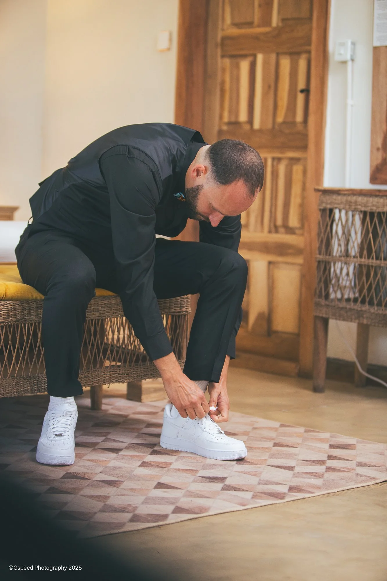 A man in black clothing tying his white sneakers inside a room with wooden doors and furniture.
