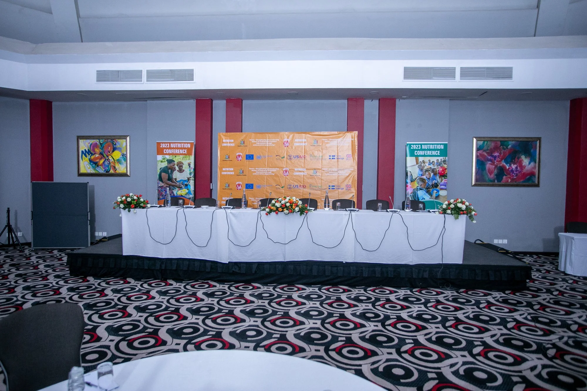 Empty conference room with a long white table, floral centerpieces, and a backdrop with event banners for the 2023 Nutrition Conference. The room has colorful abstract paintings on the walls and patterned carpet.