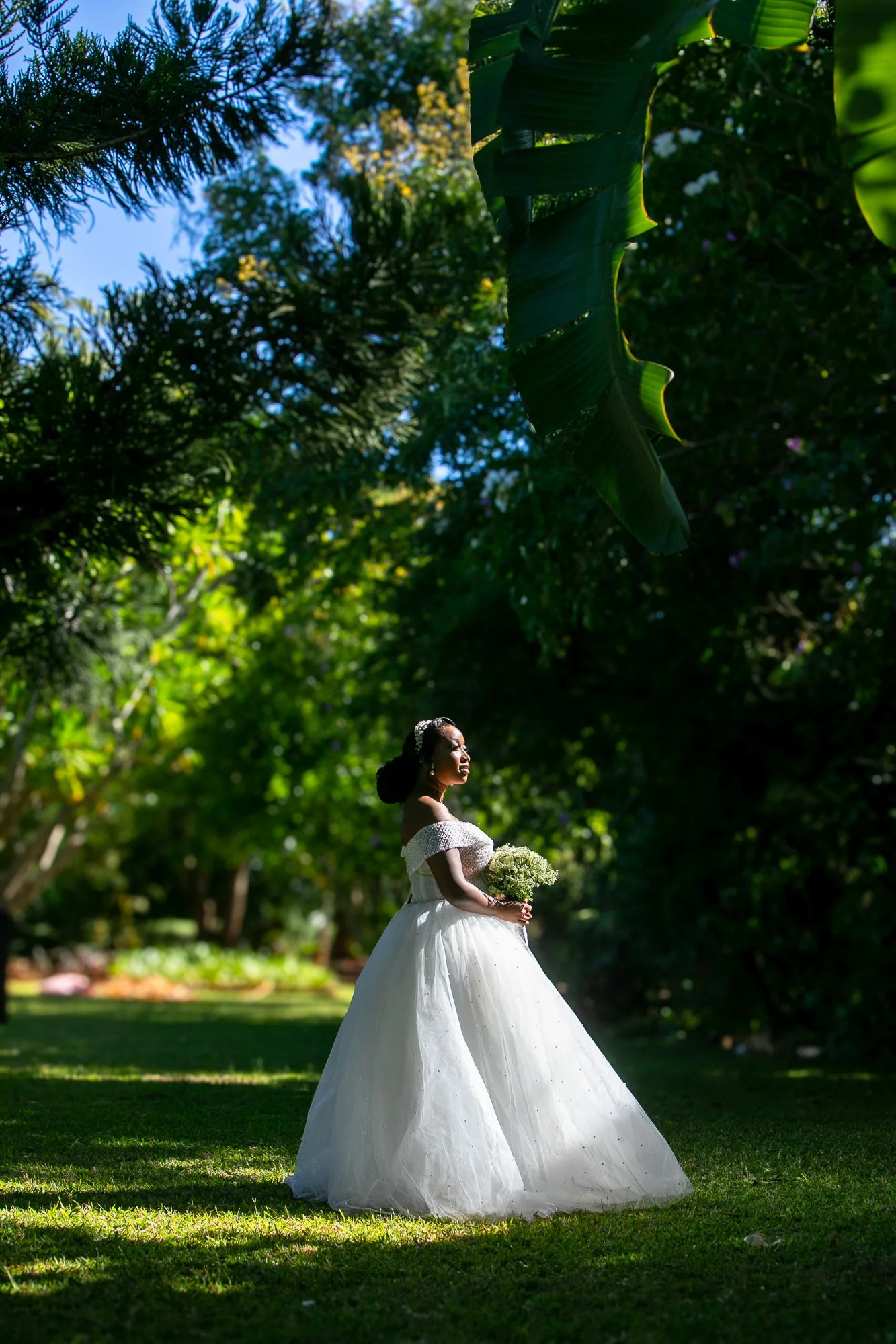 A bride in a white wedding dress holding a bouquet of flowers in a lush green outdoor garden, with trees and plants surrounding her.