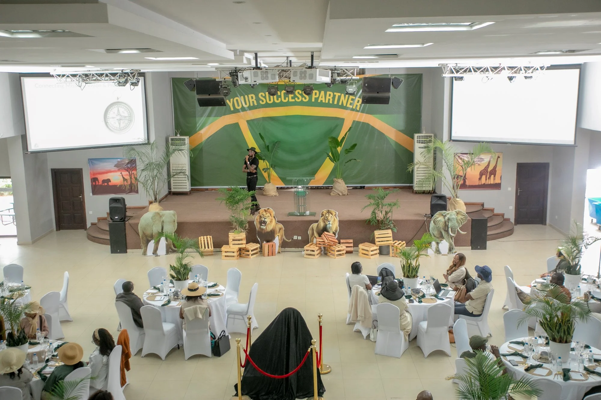 Conference hall with a stage decorated with African wildlife photos, plants, lion cutouts, and wooden crates, with audience seated at tables and a speaker on stage.