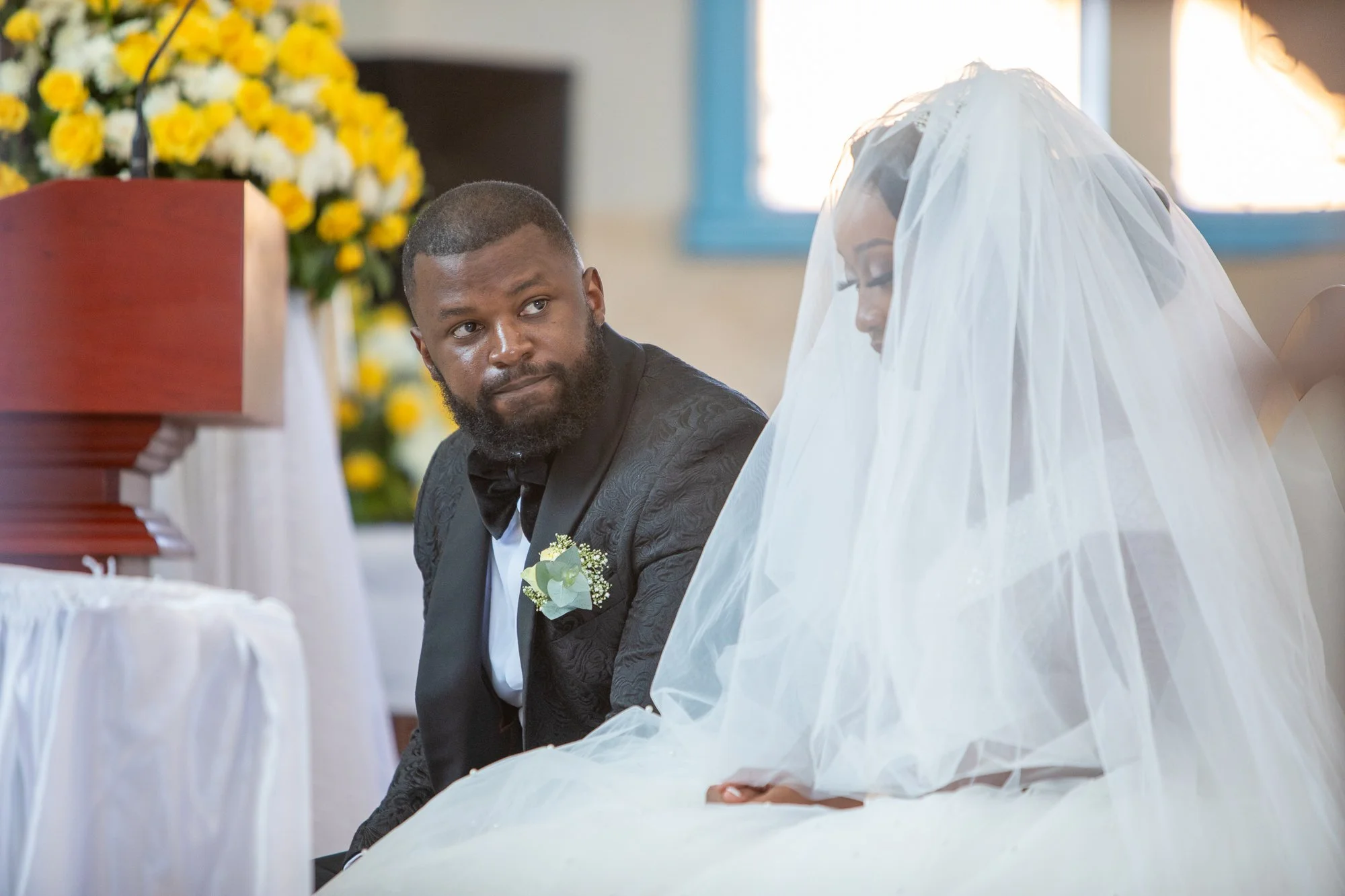 A man and a woman, dressed in formal attire with the woman wearing a white veil, are participating in a wedding ceremony. The man is looking at the woman, who is bowing her head, in a setting decorated with flowers and candles.
