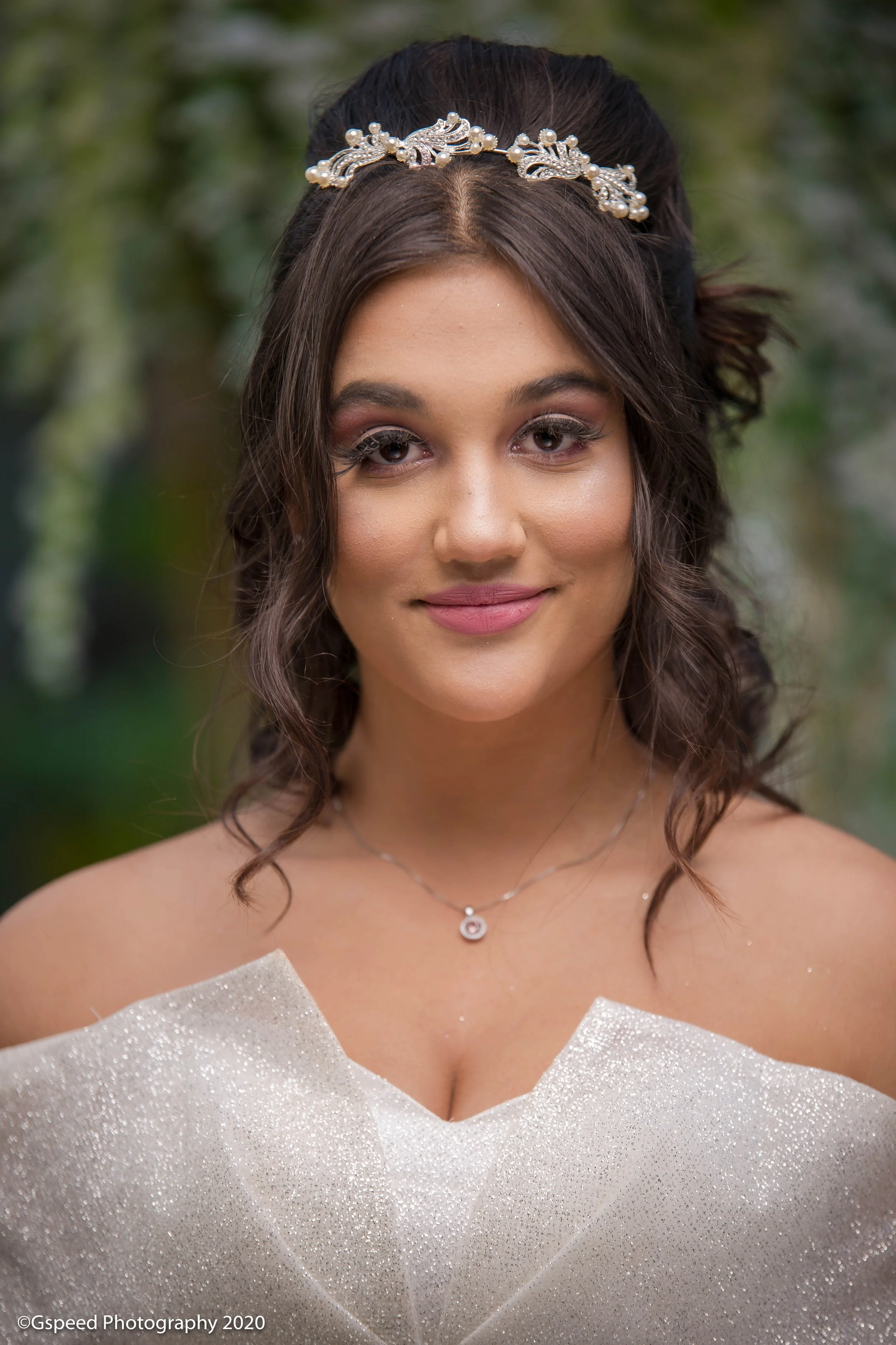 A young woman with dark wavy hair, wearing a pearl and rhinestone headband, makeup with pink lipstick and eye shadow, a delicate necklace with a small pendant, and a strapless sparkly dress, smiling outdoors with green foliage blurred in the backgrou