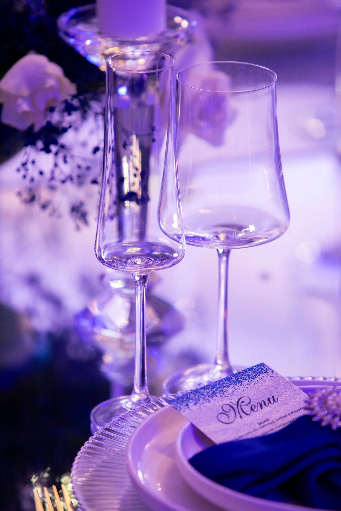 Clear champagne flutes on a decorated table with a menu, plates, and flowers, illuminated by soft purple lighting, indicating a festive event or celebration.