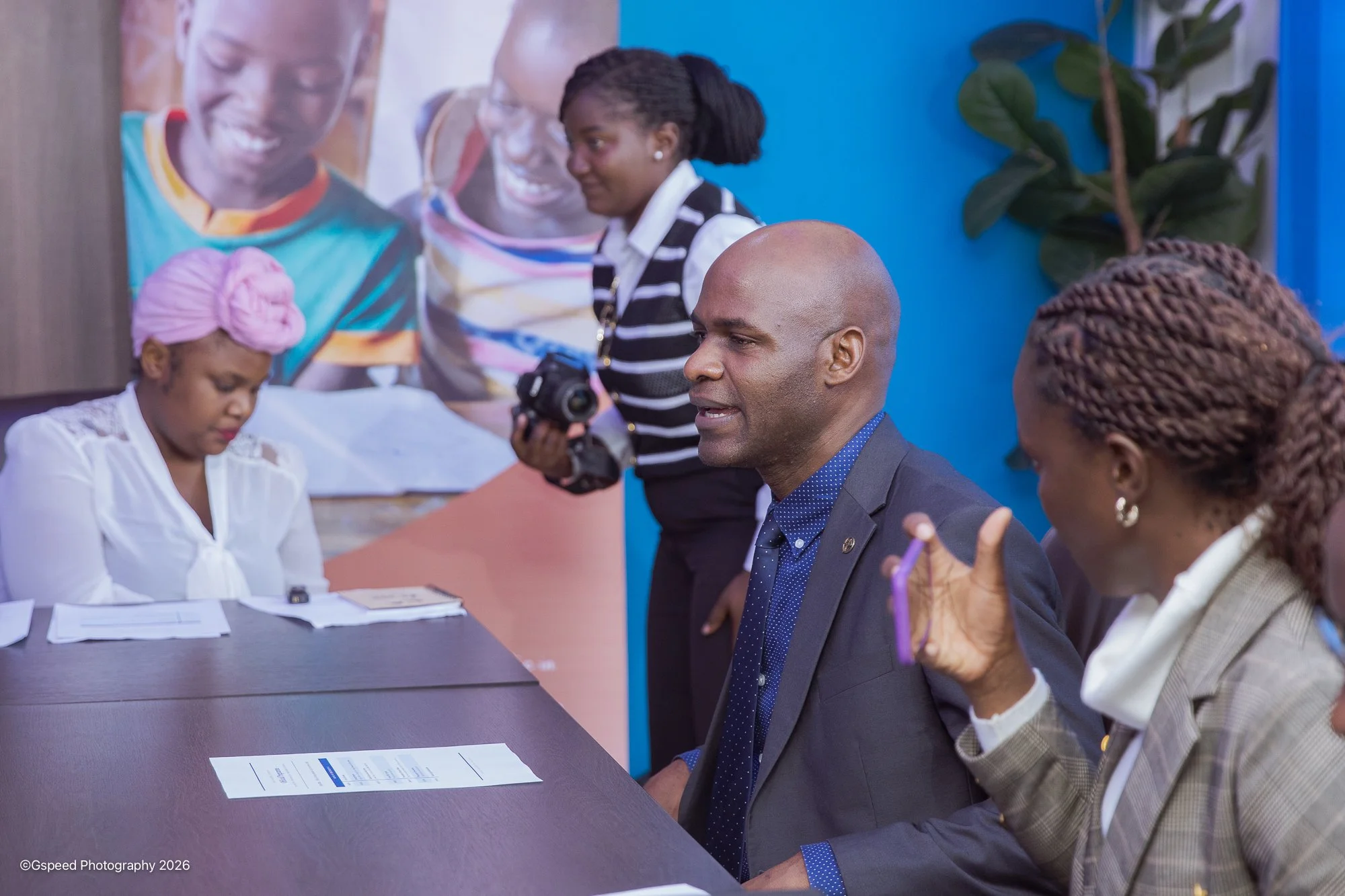 A group of four diverse individuals, three women and one man, engaged in a discussion at a table in a professional setting. The man, seated, is speaking while the women are listening, with one woman taking notes. In the background, a woman is standin
