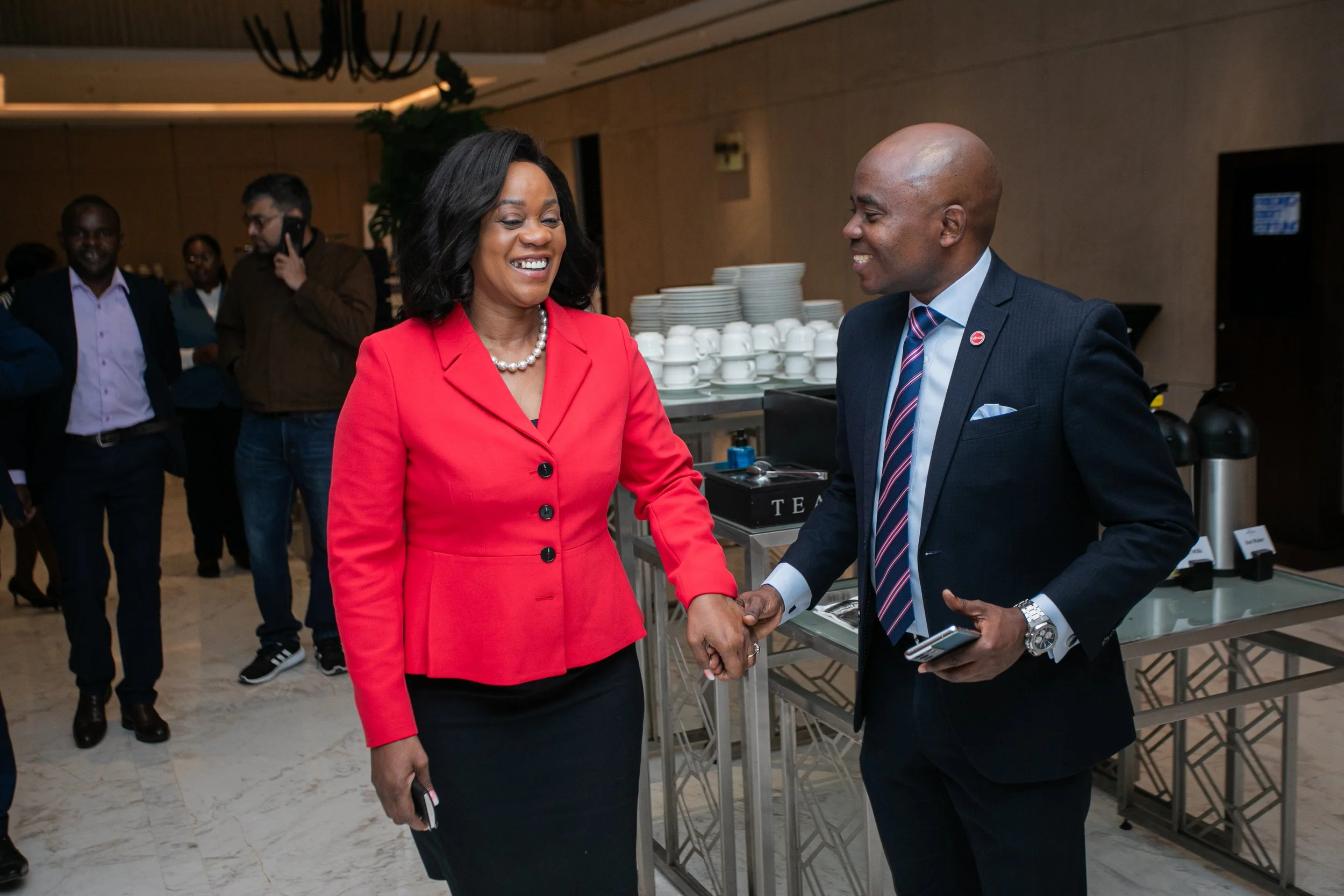 A woman in a bright red blazer and black skirt shaking hands with a man in a dark suit and striped tie. They are smiling at each other in a professional setting with a group of people in the background and a table with stacked dishes.