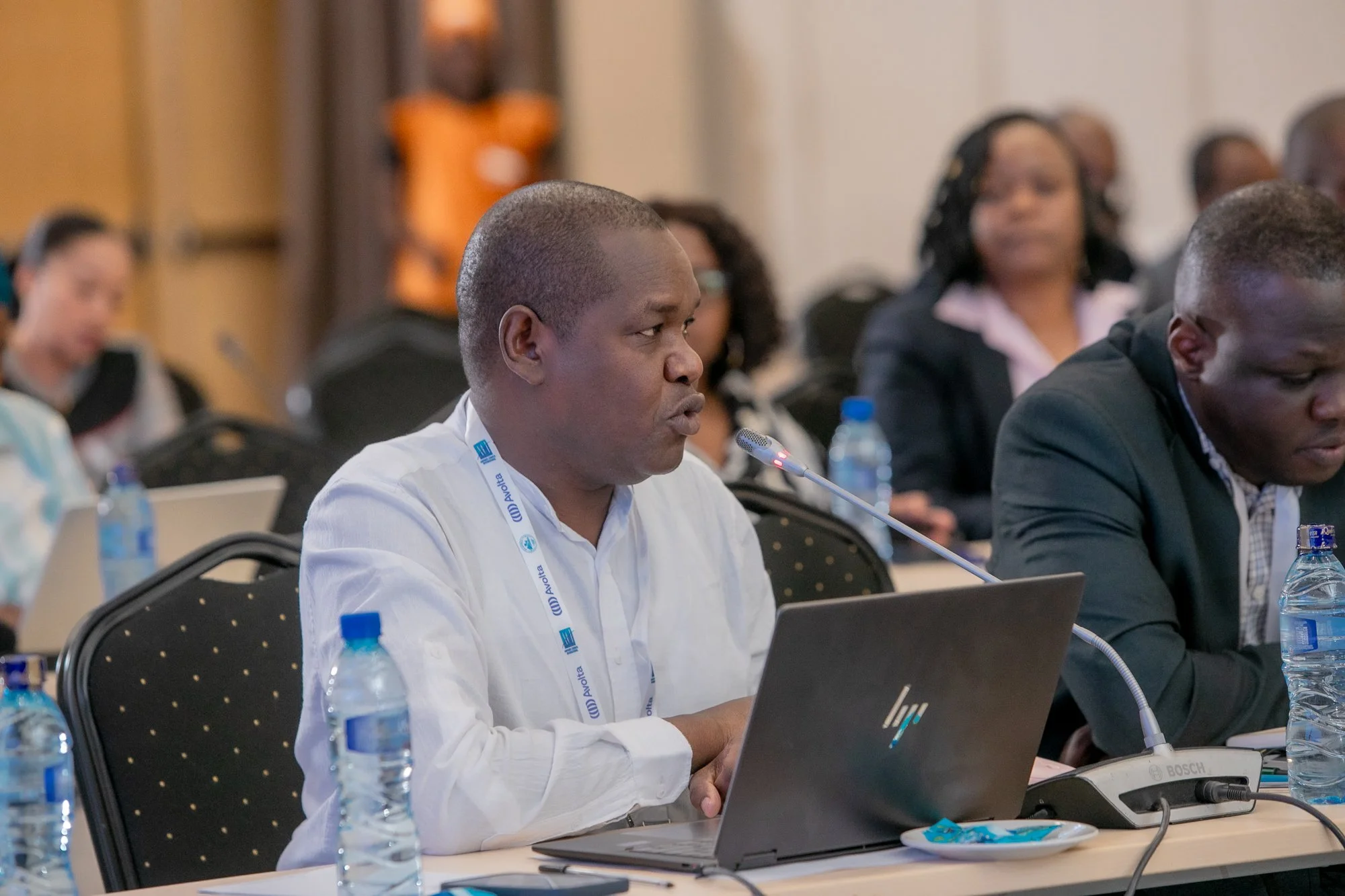 Man in white shirt attending a conference, sitting at a desk with a laptop, microphone, and water bottles, with other attendees visible in the background.