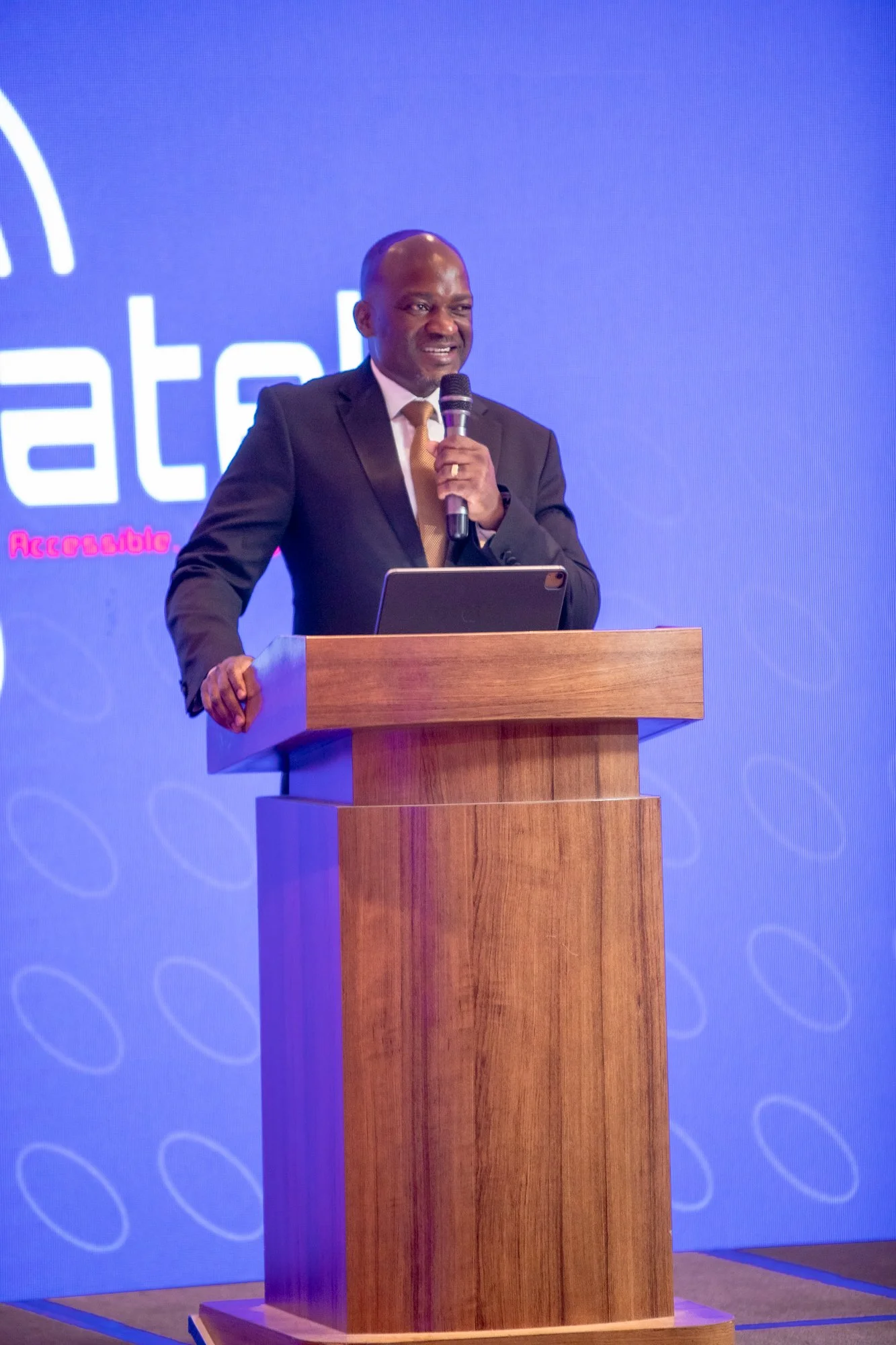 A man in a suit and tie is speaking into a microphone at a podium during a presentation or conference. The background features a blue screen with a logo and text partially visible.