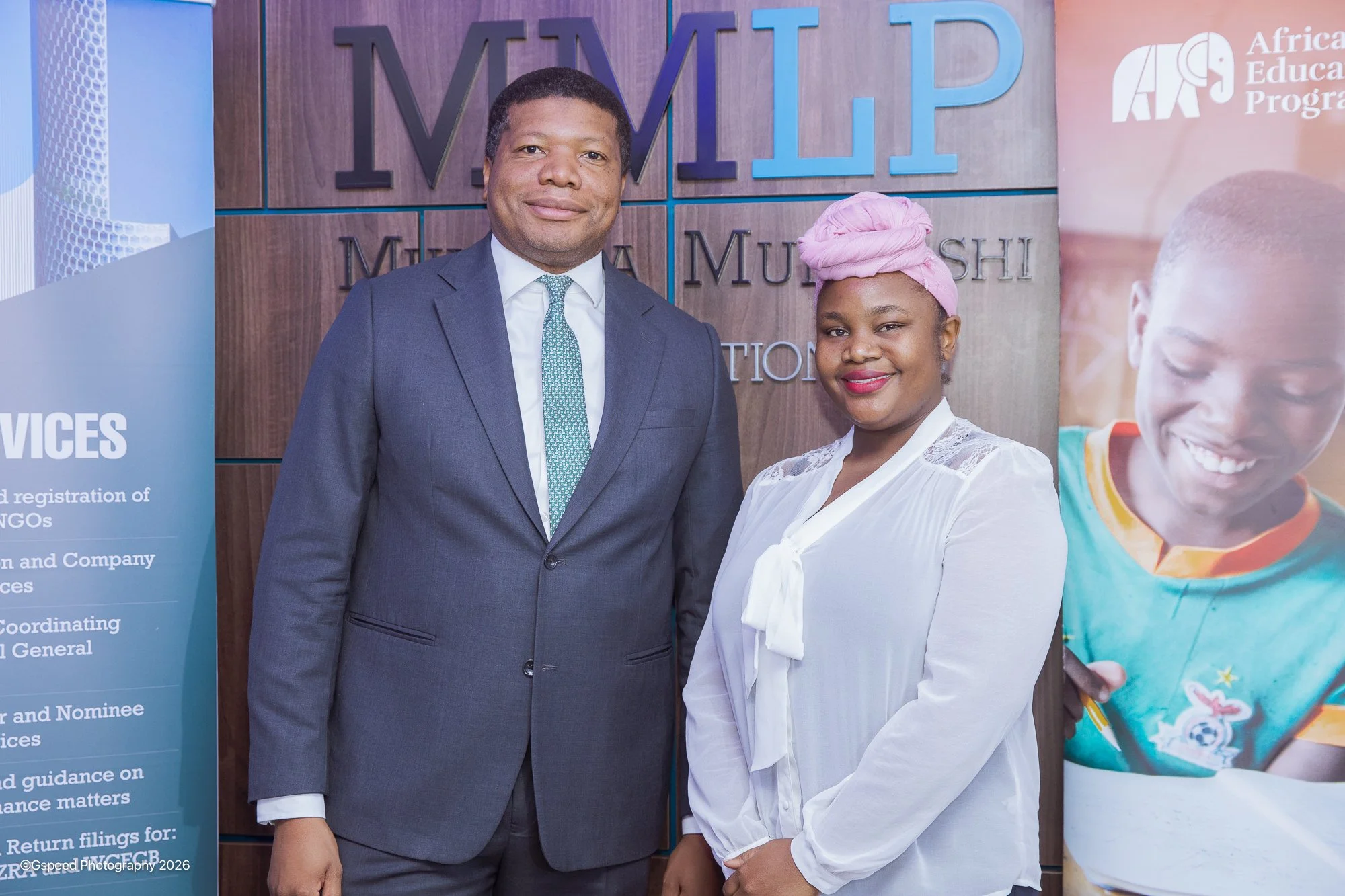 A man in a gray suit and a woman in a white blouse with a pink headwrap standing in front of a wooden wall with large letters that spell MWMTP. The woman is smiling, and the man has a neutral expression.