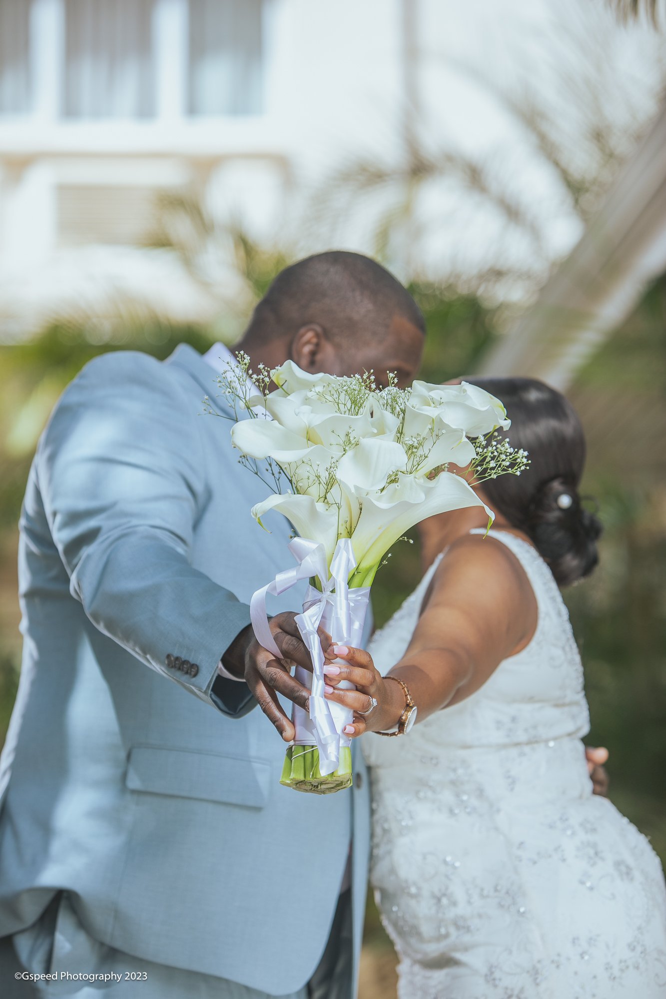 A couple dressed in wedding attire holding a bouquet of white calla lilies and baby’s breath during an outdoor ceremony.