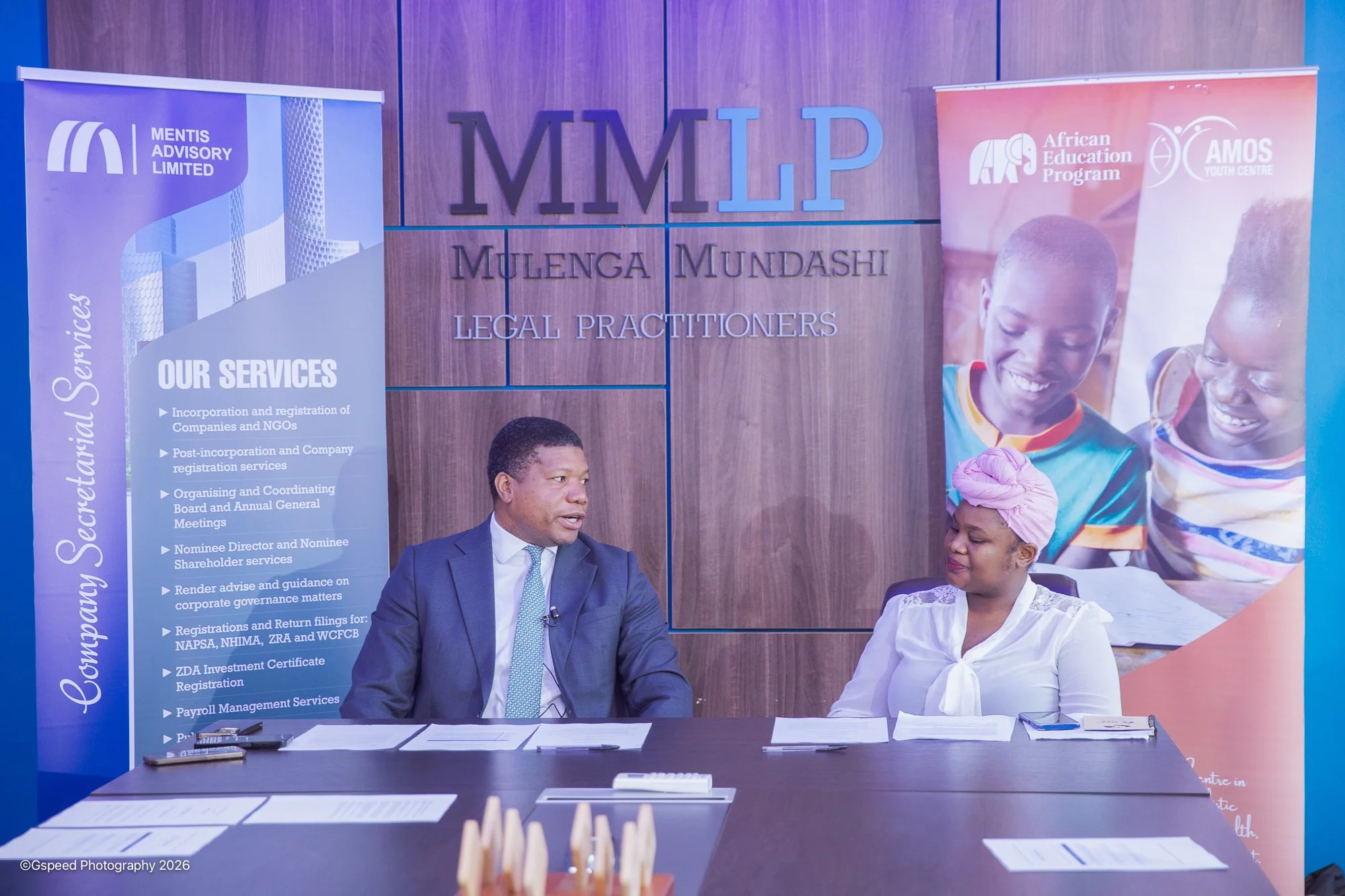 A man in a suit and a woman in a white blouse sit at a black table with papers and pens, in front of display banners for Mulenga Mundashi Legal Practitioners and the African Education Program Youth Centre. The background shows a wooden wall with the 