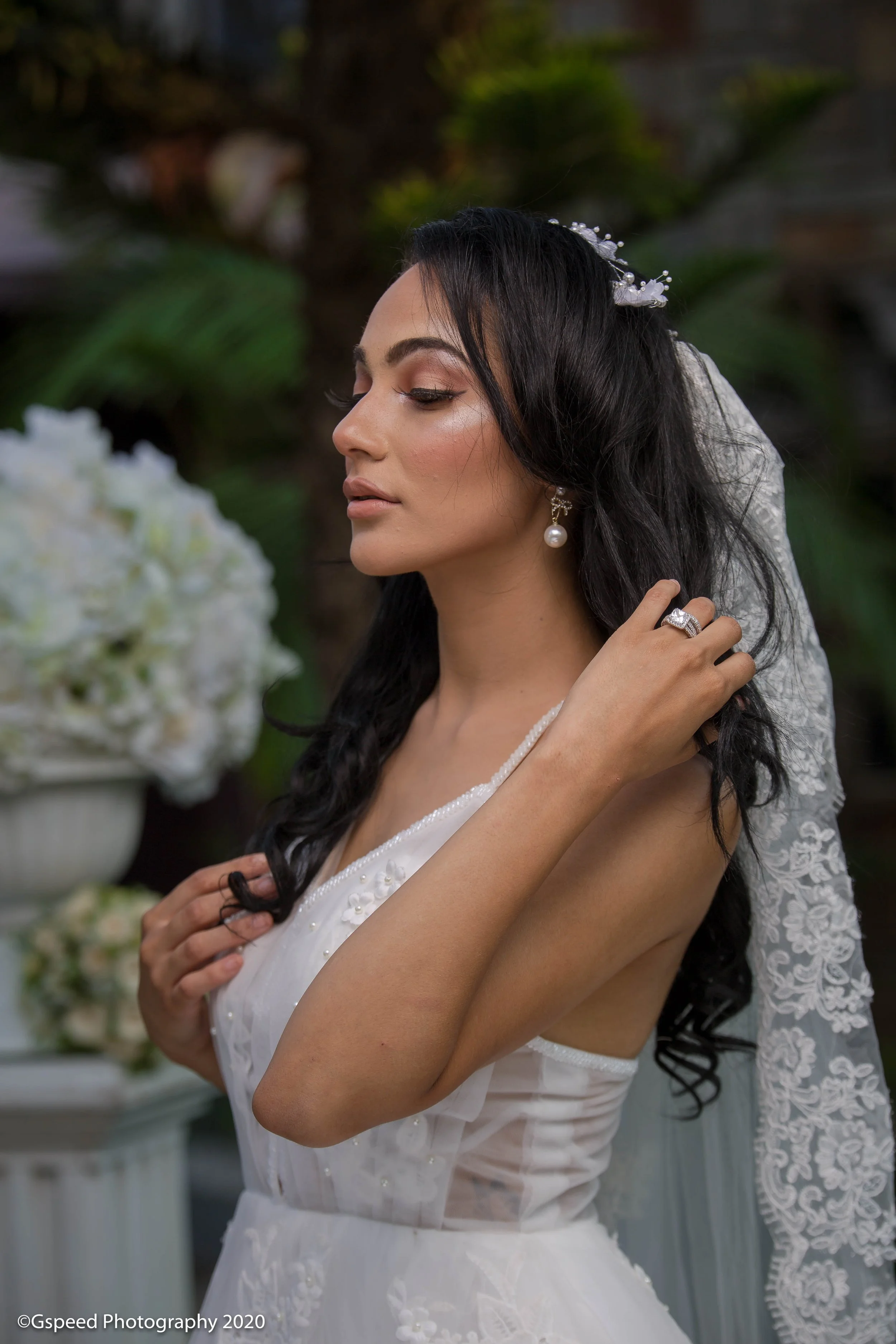 A woman with dark, wavy hair and pearl earrings in a white wedding dress, adjusting her hair with her eyes closed, in a garden setting with flowers and greenery in the background.