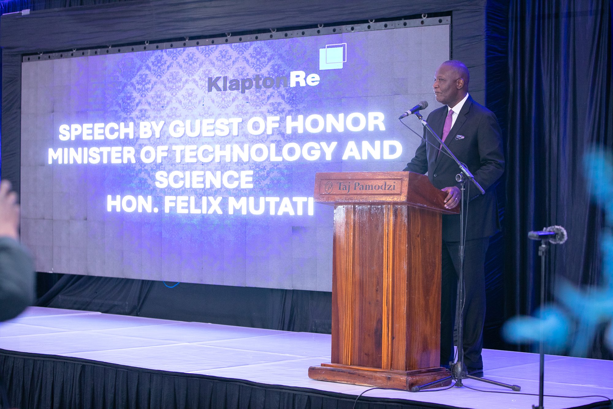 A man dressed in a dark suit, white shirt, and purple tie stands at a wooden podium with a microphone, giving a speech at a formal event. Behind him, a large screen displays text mentioning a speech by Hon. Felix Mutati, Minister of Technology and Sc