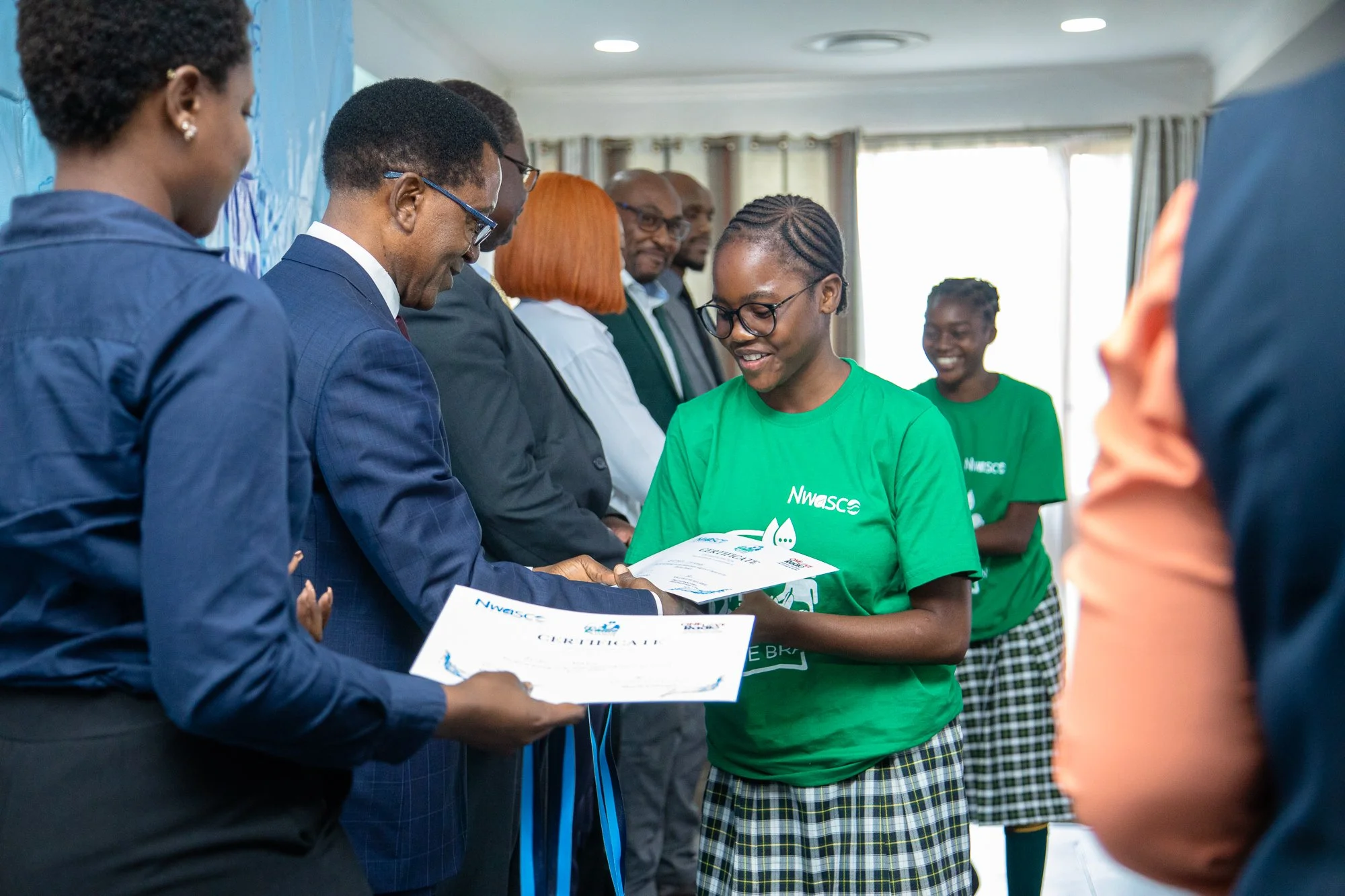A young woman in a green t-shirt receiving a certificate from a man in a suit at an indoor award ceremony, with other smiling people in the background.