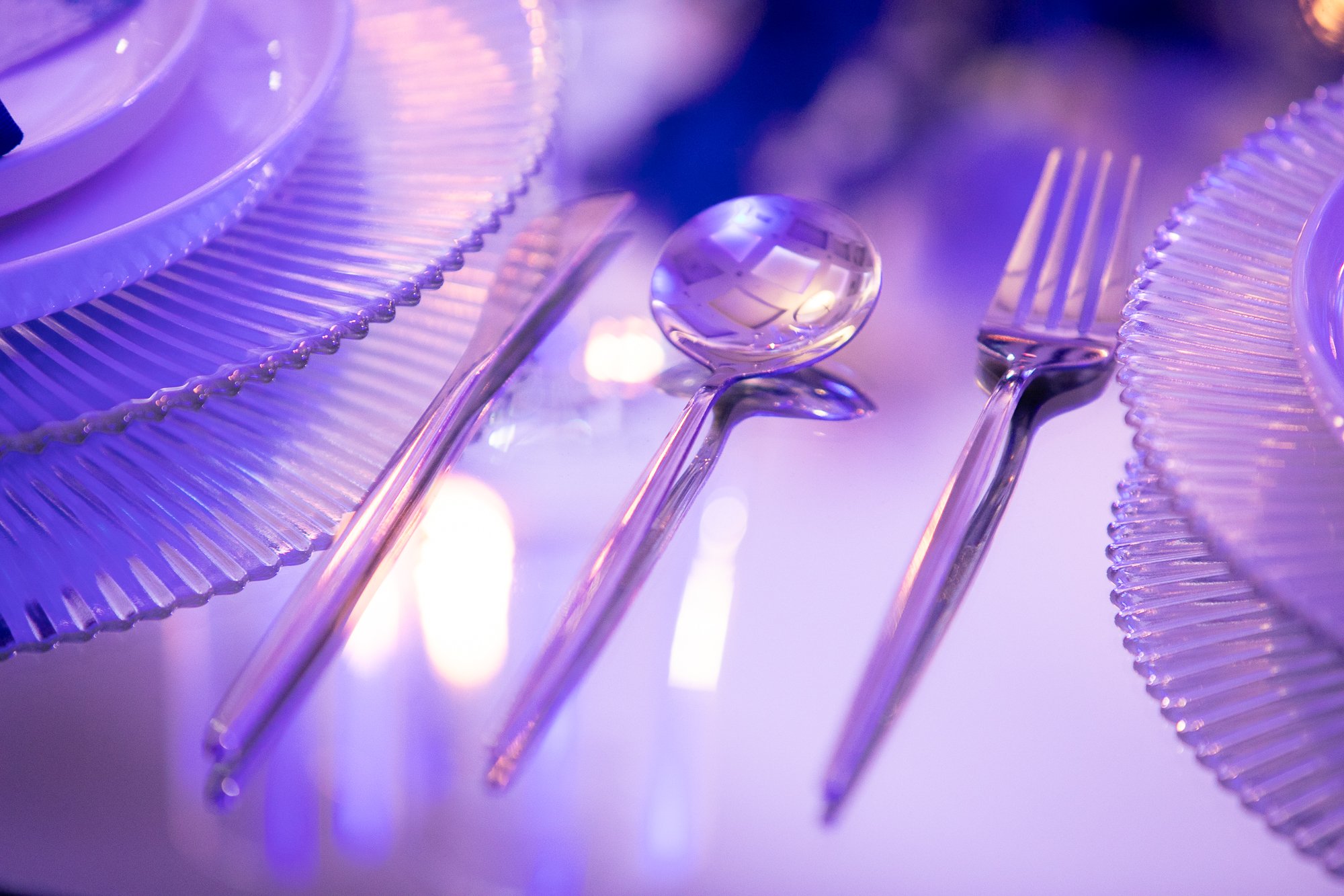 Various transparent glass cutlery including forks, knives, and spoons with prismatic ridges, illuminated with purple lighting, reflecting light and creating a shiny, iridescent appearance.